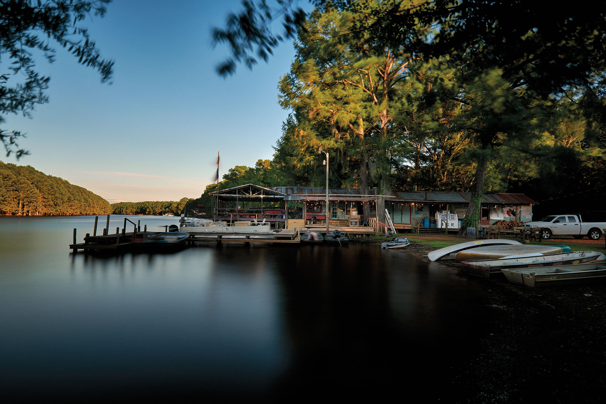 A store with a large dock on a smooth lake next to a rolling green hill