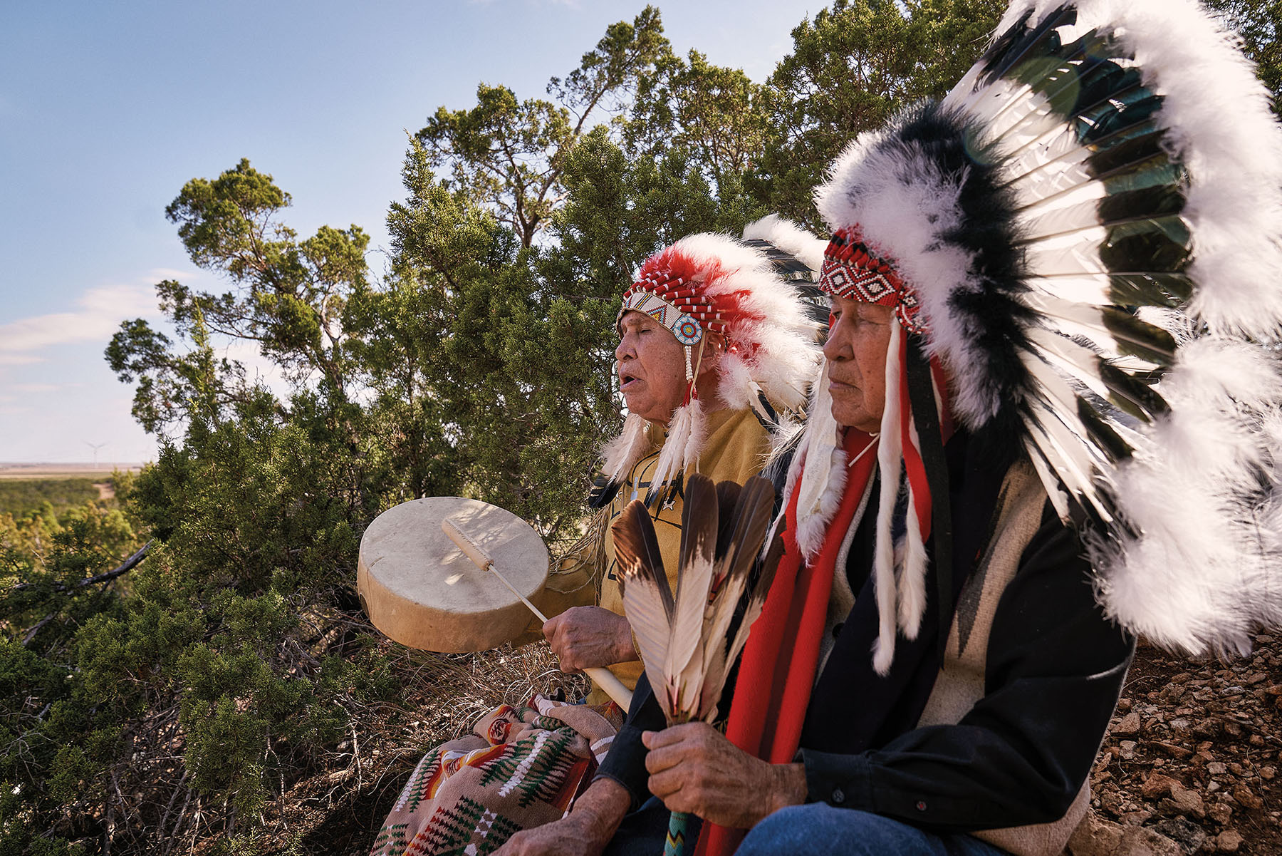 Two men dressed in traditional Native American attire sit on a rock playing a drum under blue sky