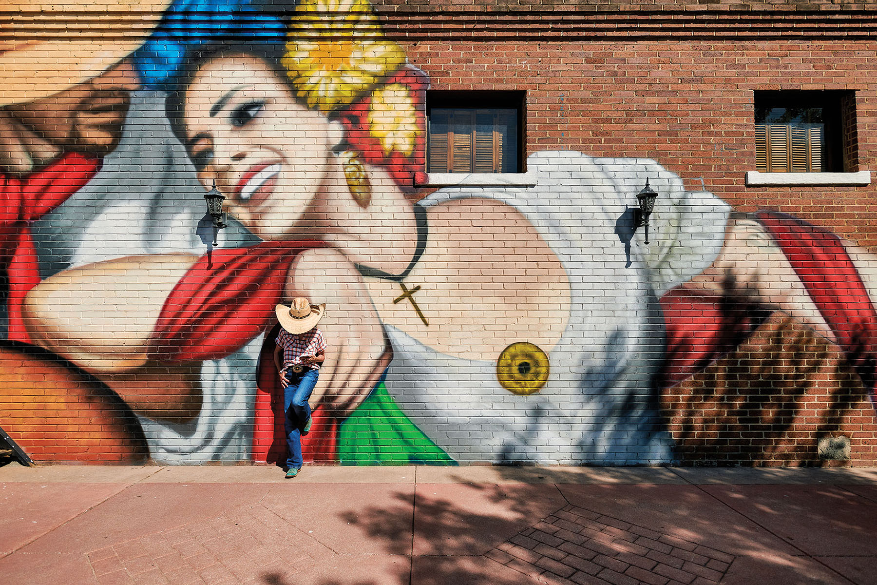 A person in a cowboy hat leans against a mural of two people dancing, painted on a brick wall