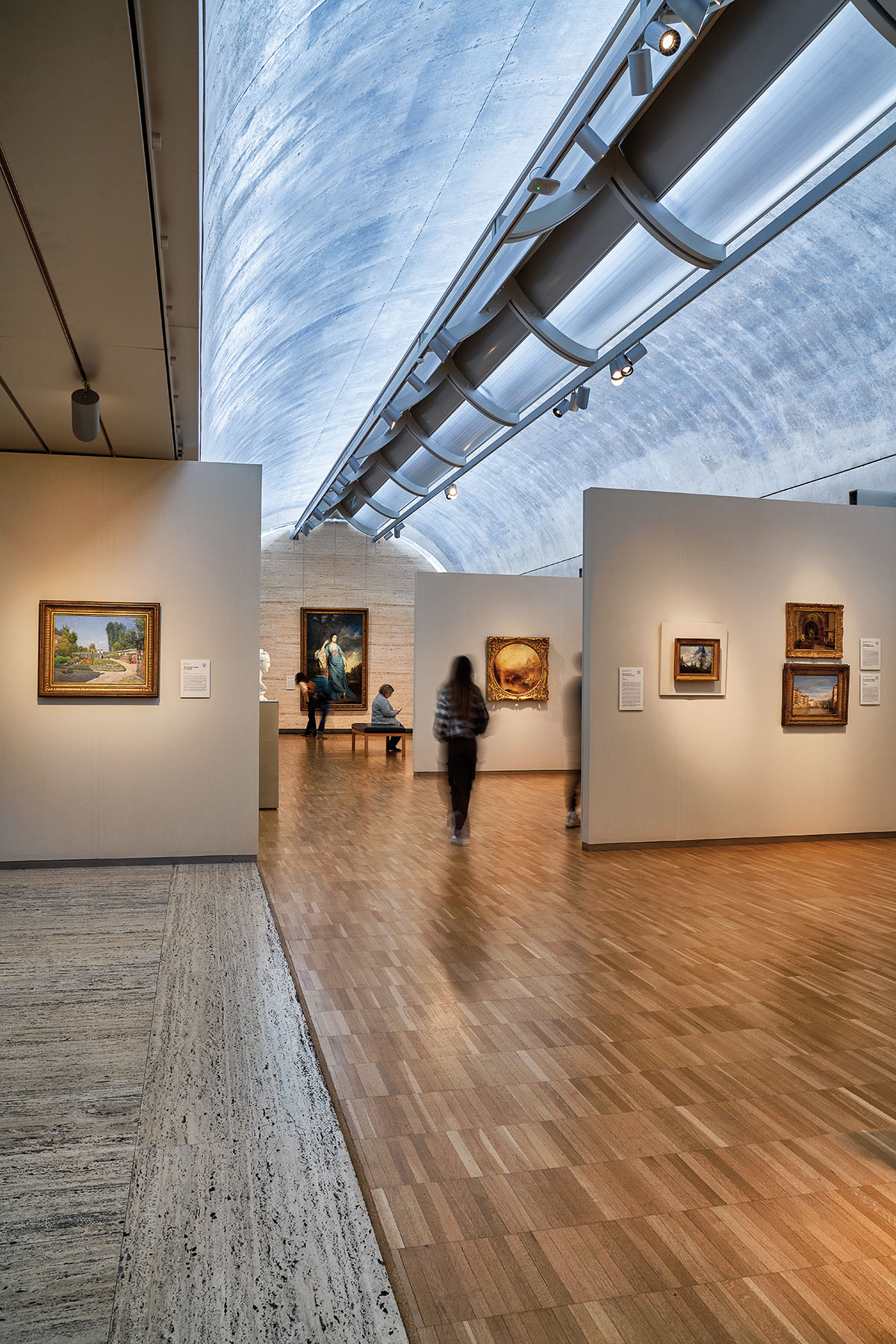 A blurred figure walks between several walls in a gallery with a diffused skylight overhead