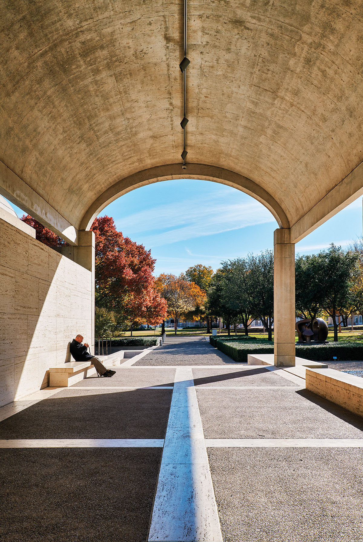 A man rests on a bench beneath a curved concrete dome in front of brightly colored trees