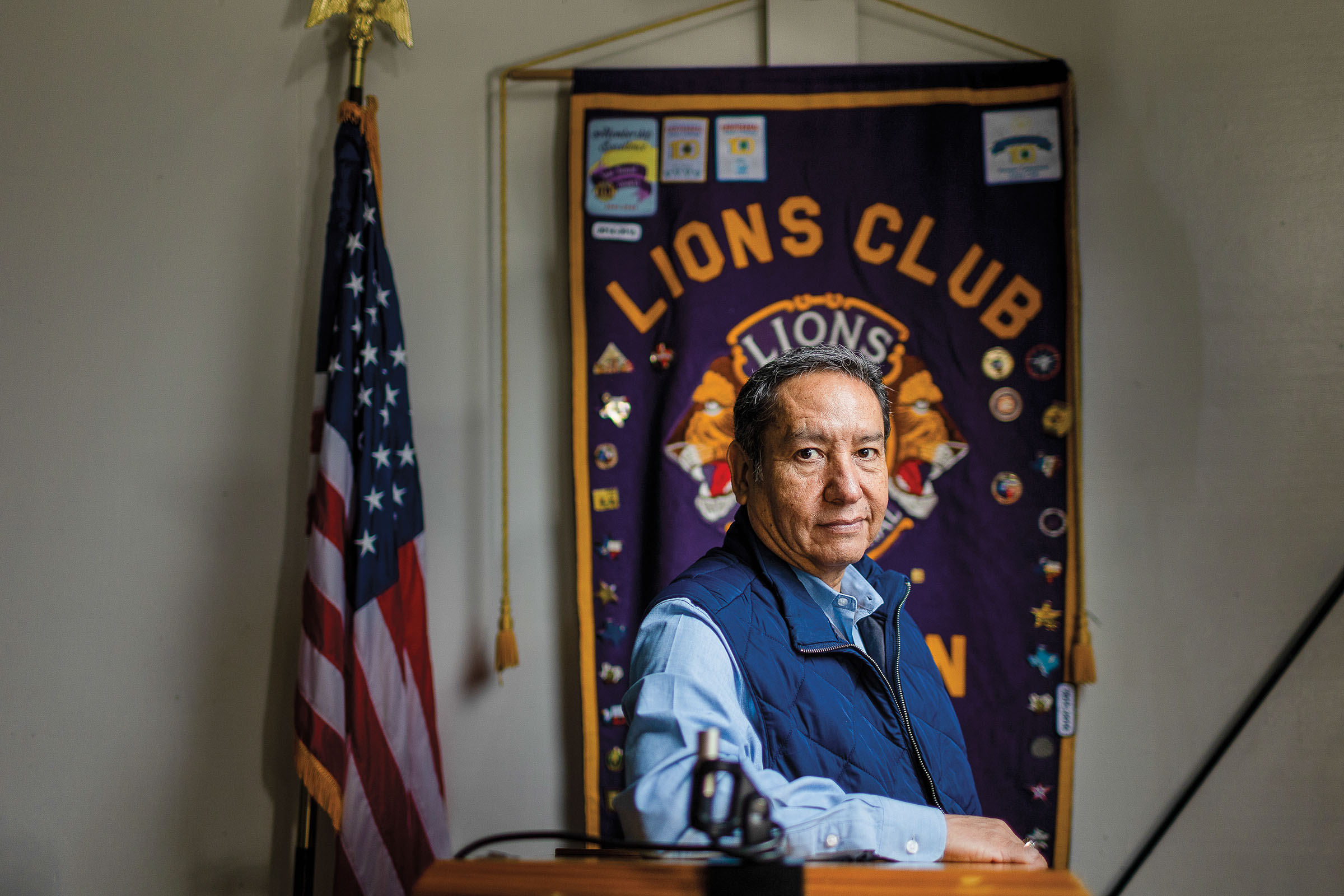 A man in a blue vest sits at a desk beneath a purple banner reading "Lions Club"