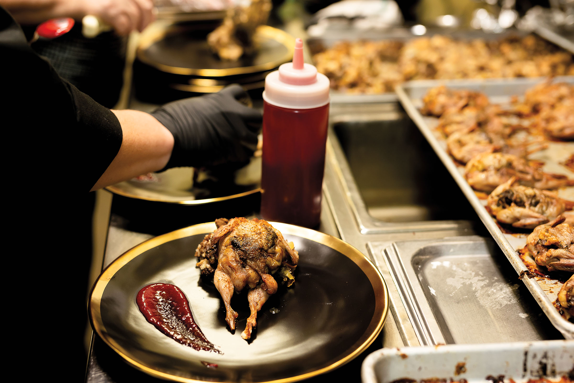 A golden brown roasted quail sits on a shiny plate next to a bright red sauce