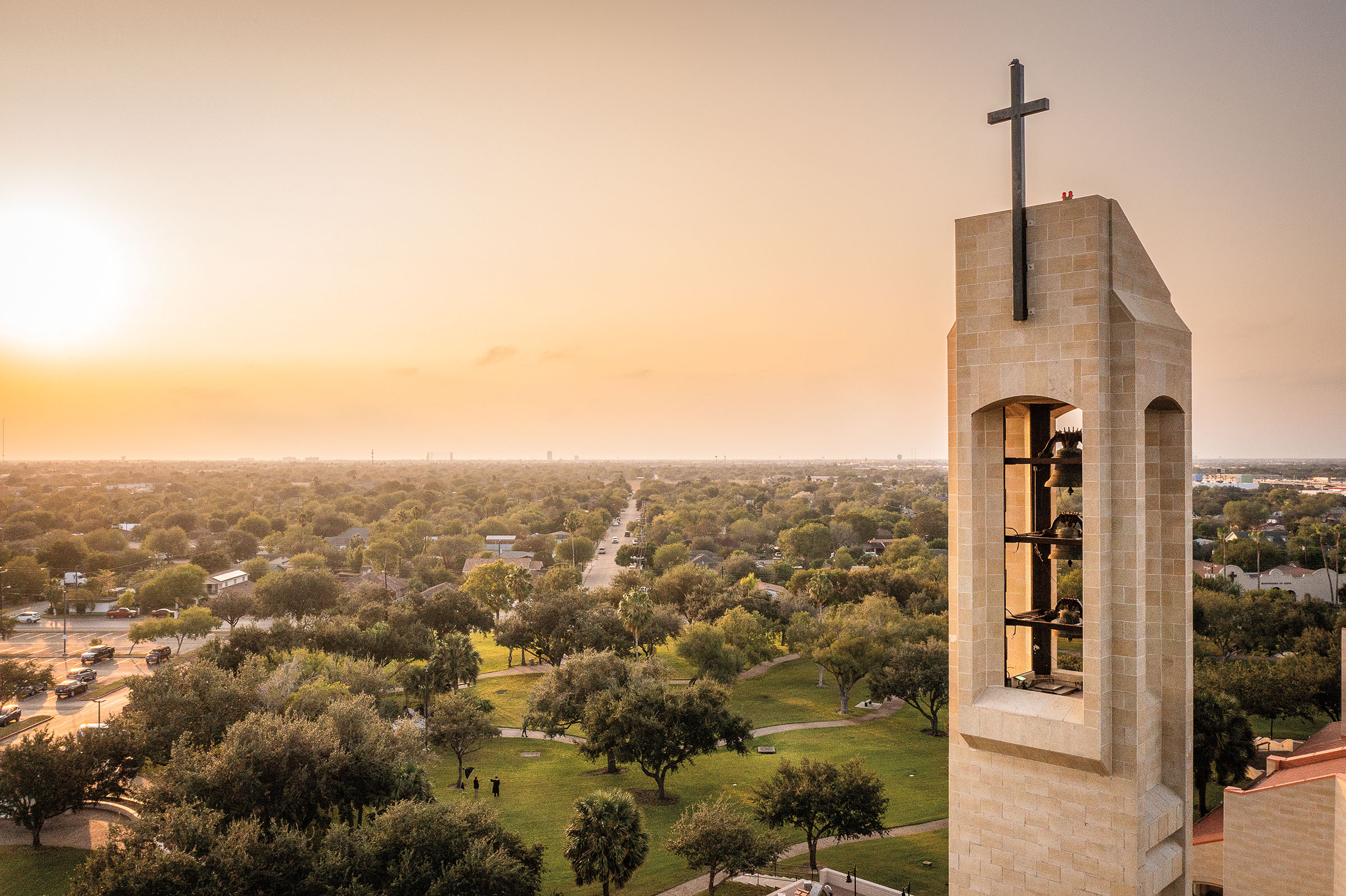 A view of a concrete church steeple with a black cross overlooking a flat green landscape with a rising sun