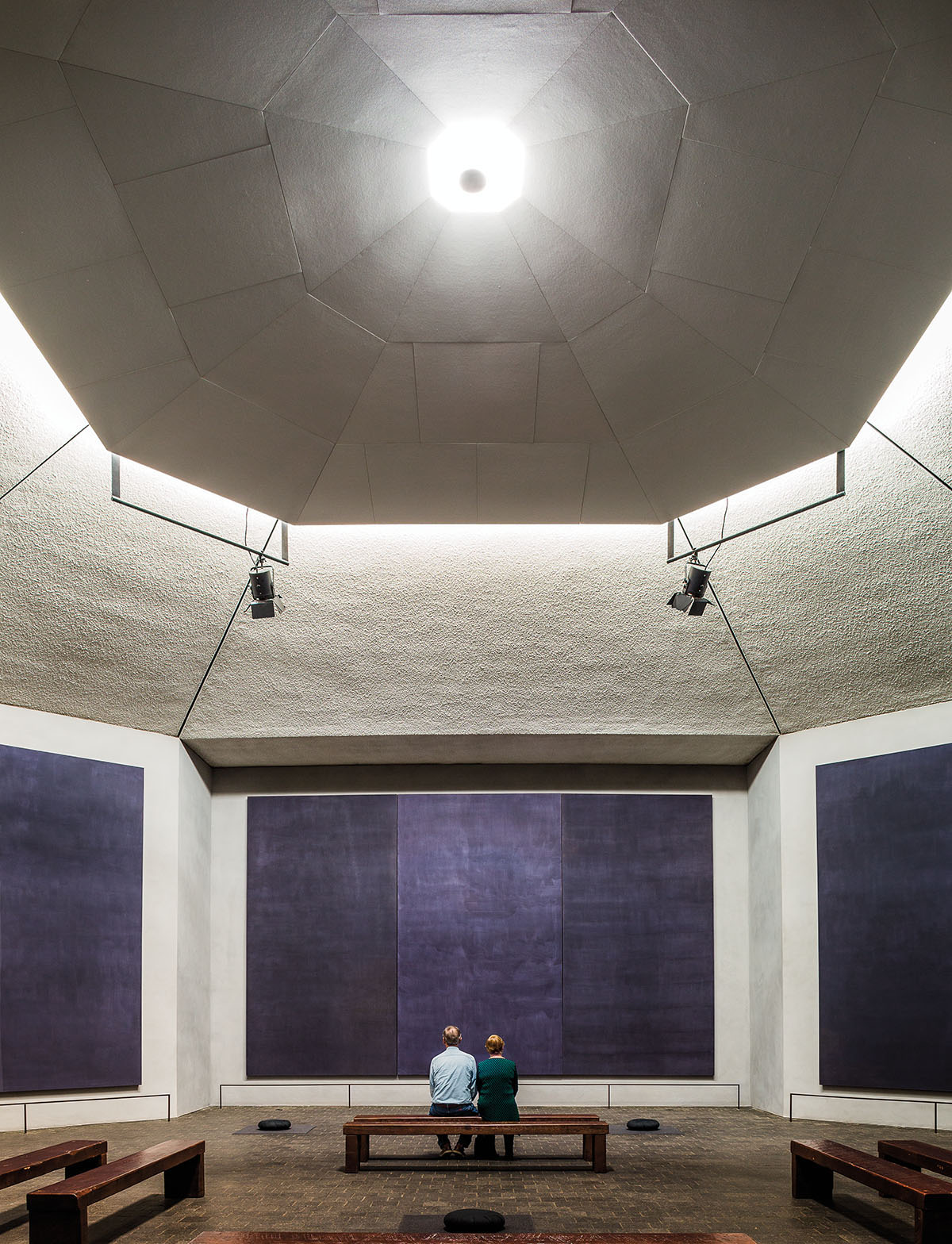 Two people sit on a wooden bench beneath a stark lit rotunda