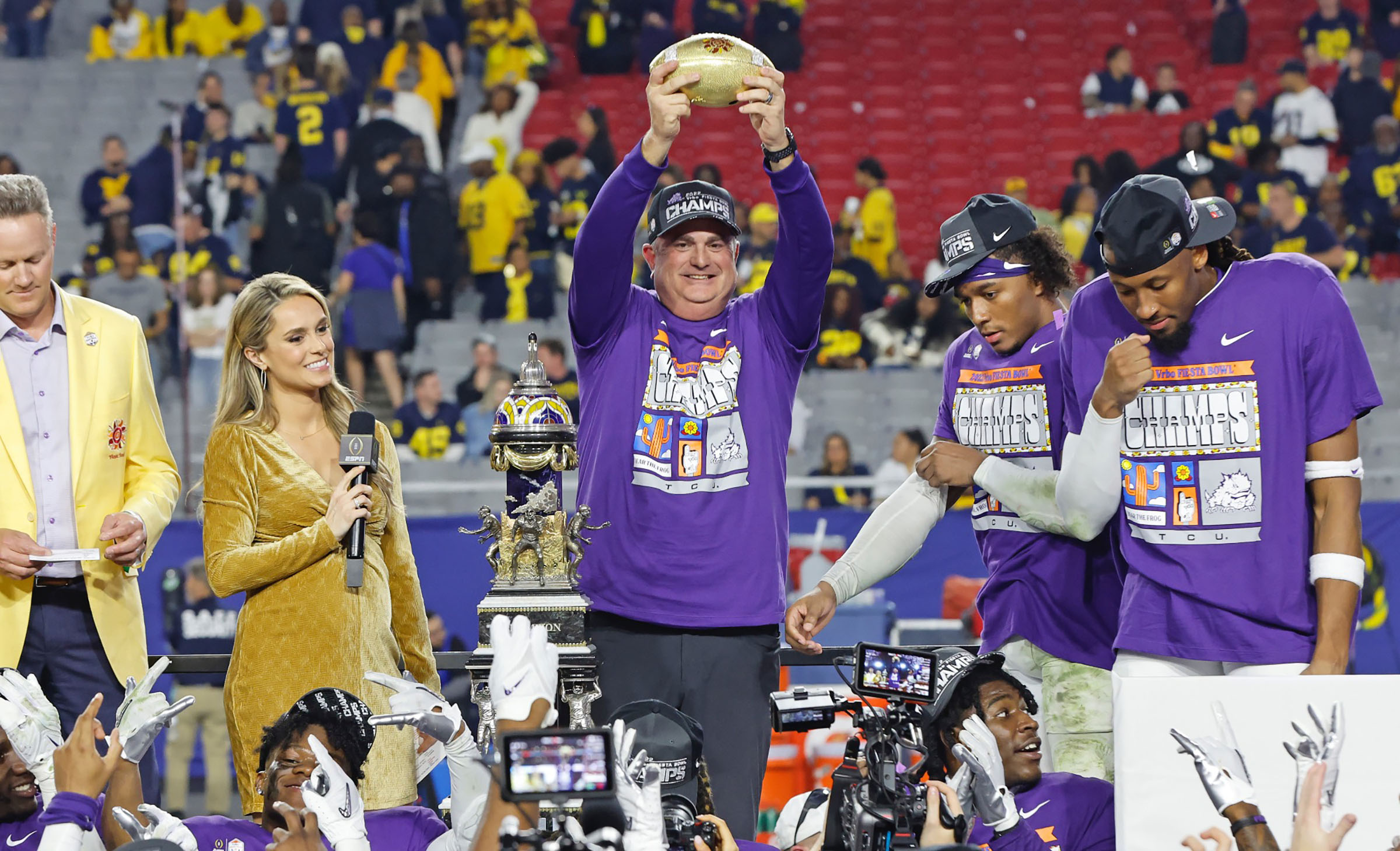 A man in a purple shirt reading "Champs" holds a golden football trophy above his head while players and spectators look on