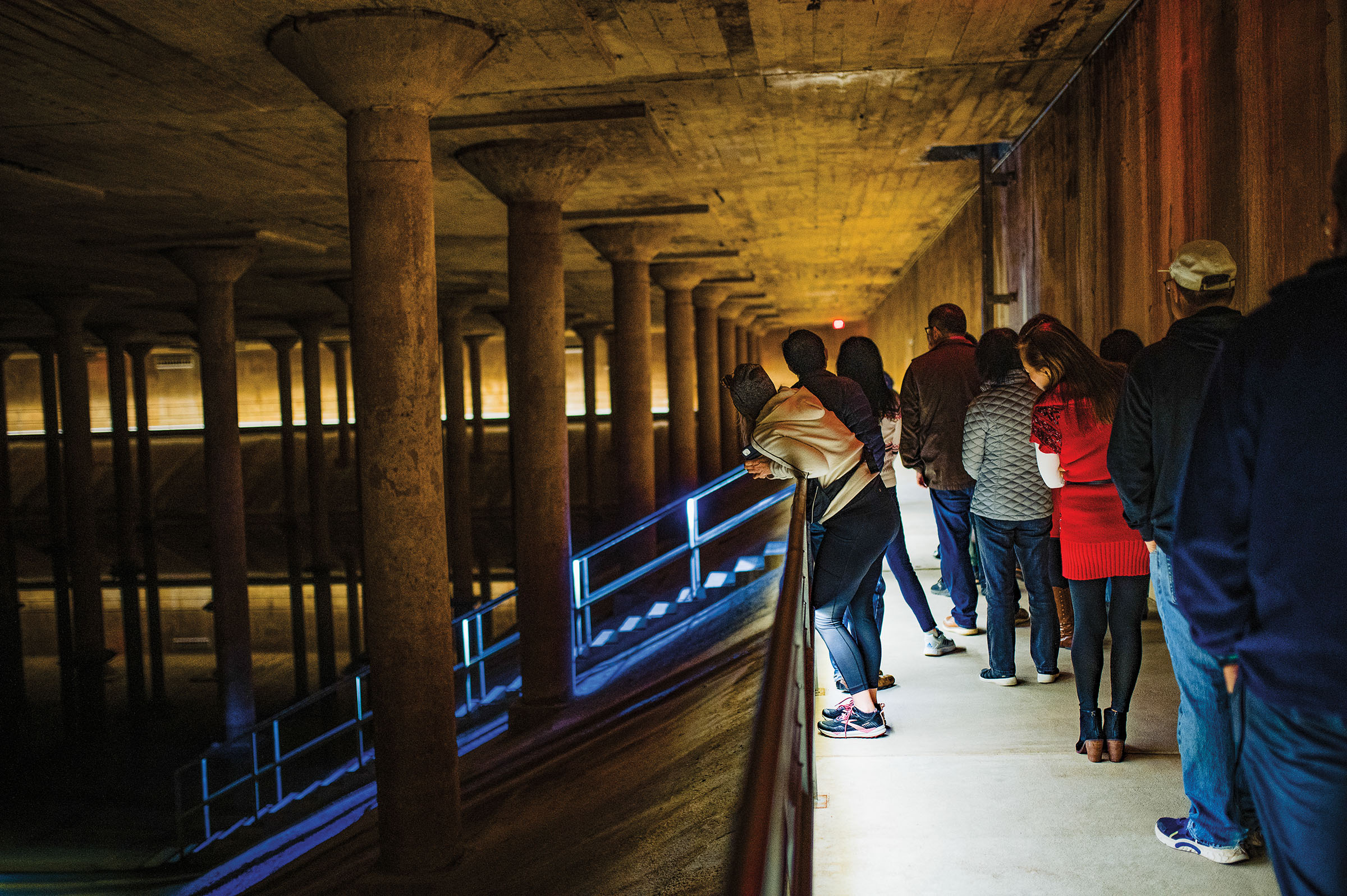 A group of people stand on a lit bridge looking into a dark cistern