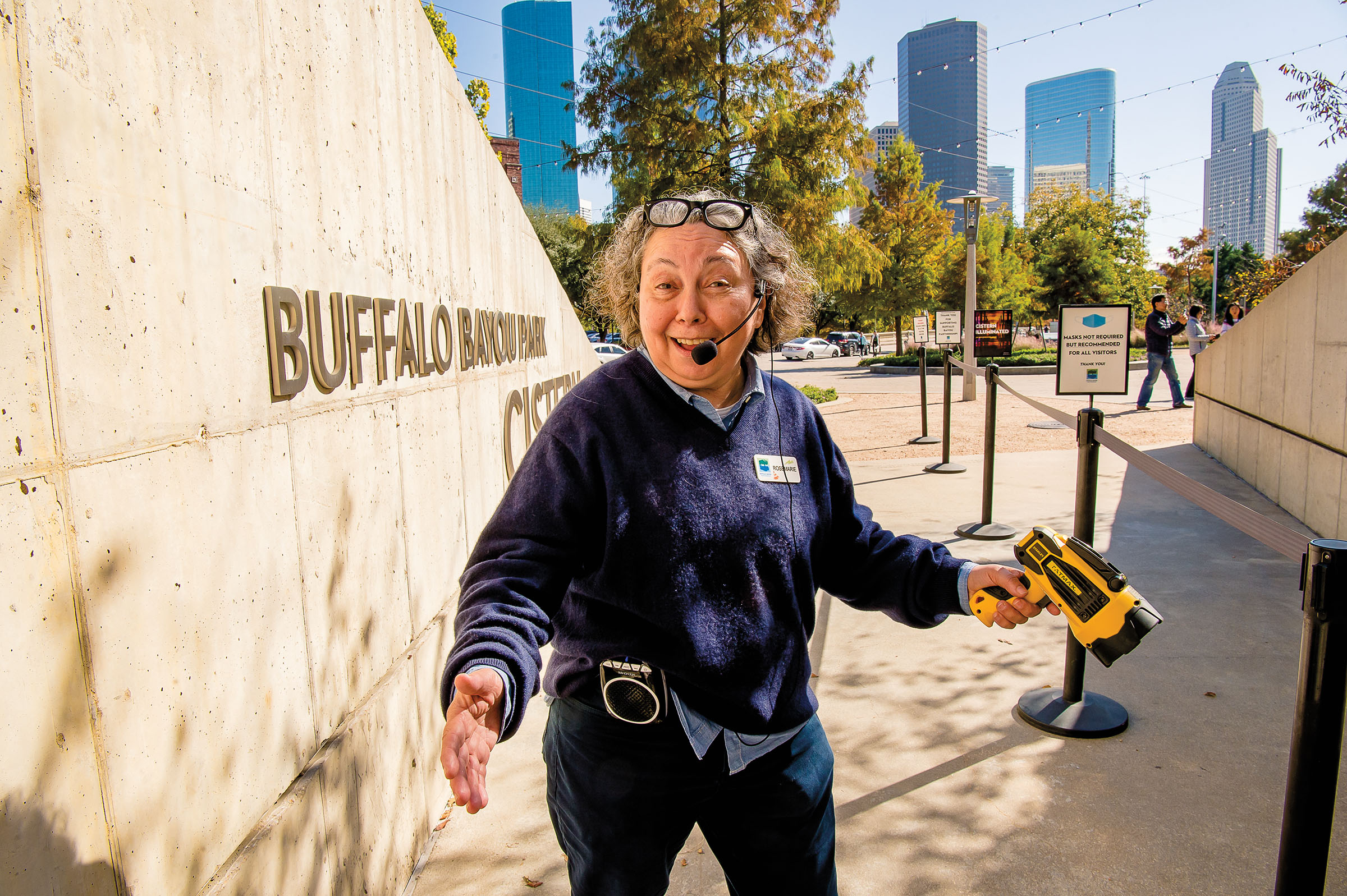 A woman in a blue sweatshirt wearing a microphone headset stands enthusiastically next to a concrete sign reading "Buffalo Bayou"
