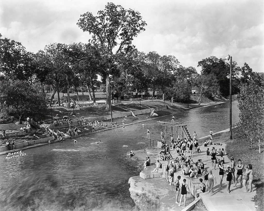 A group of people enjoy the cool water of Barton Springs Pool under green trees in a black and white photograph
