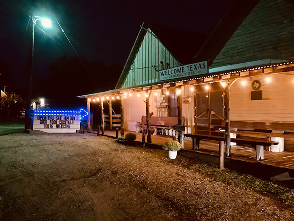 The exterior of Welcome General Store at night, with a string of lights lit up along the overhanging roof of the patio.