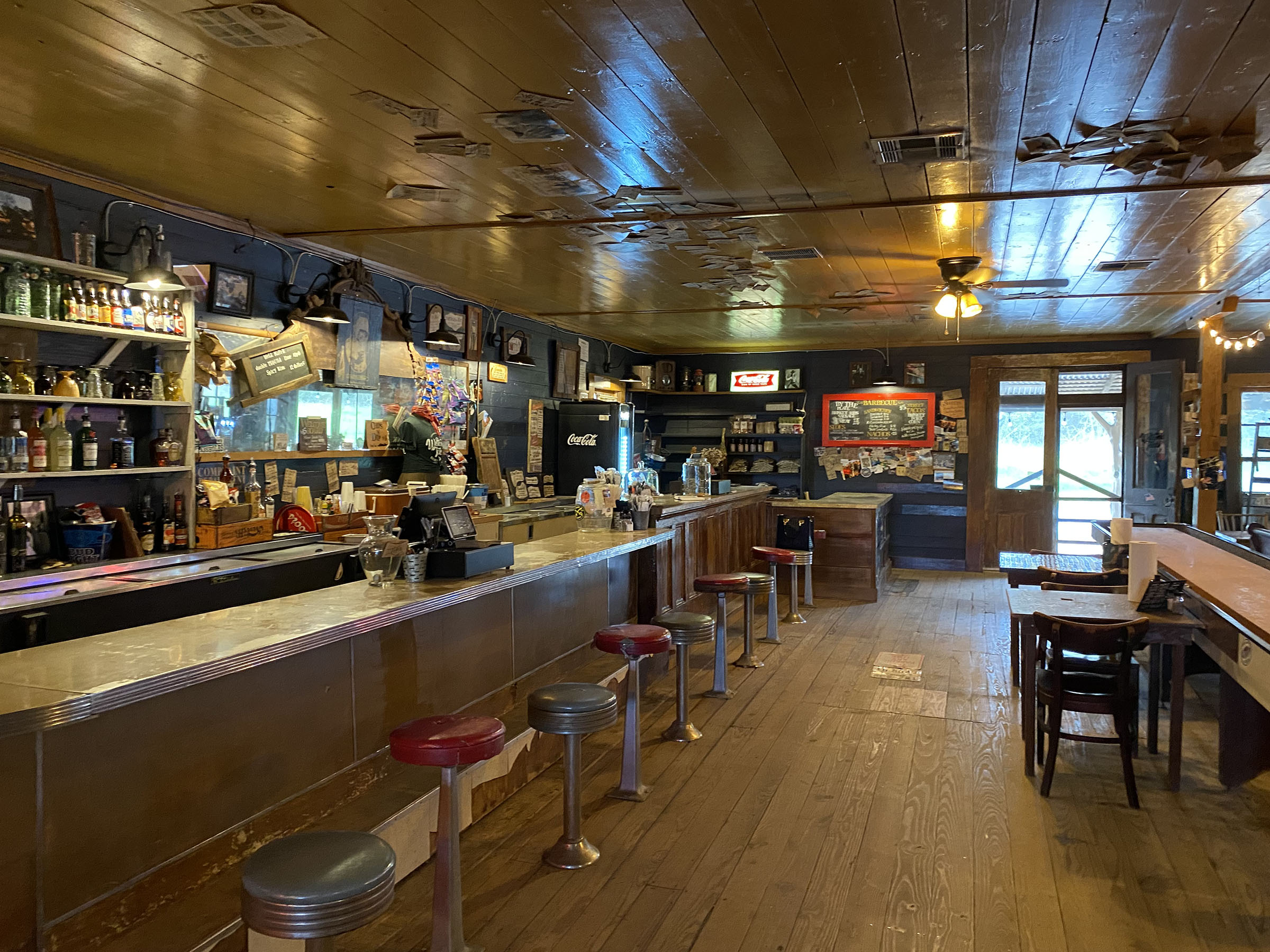 The interior of Welcome General Store shows an extended bar with bar stools, neon signs, and freshly restored wood floors. 