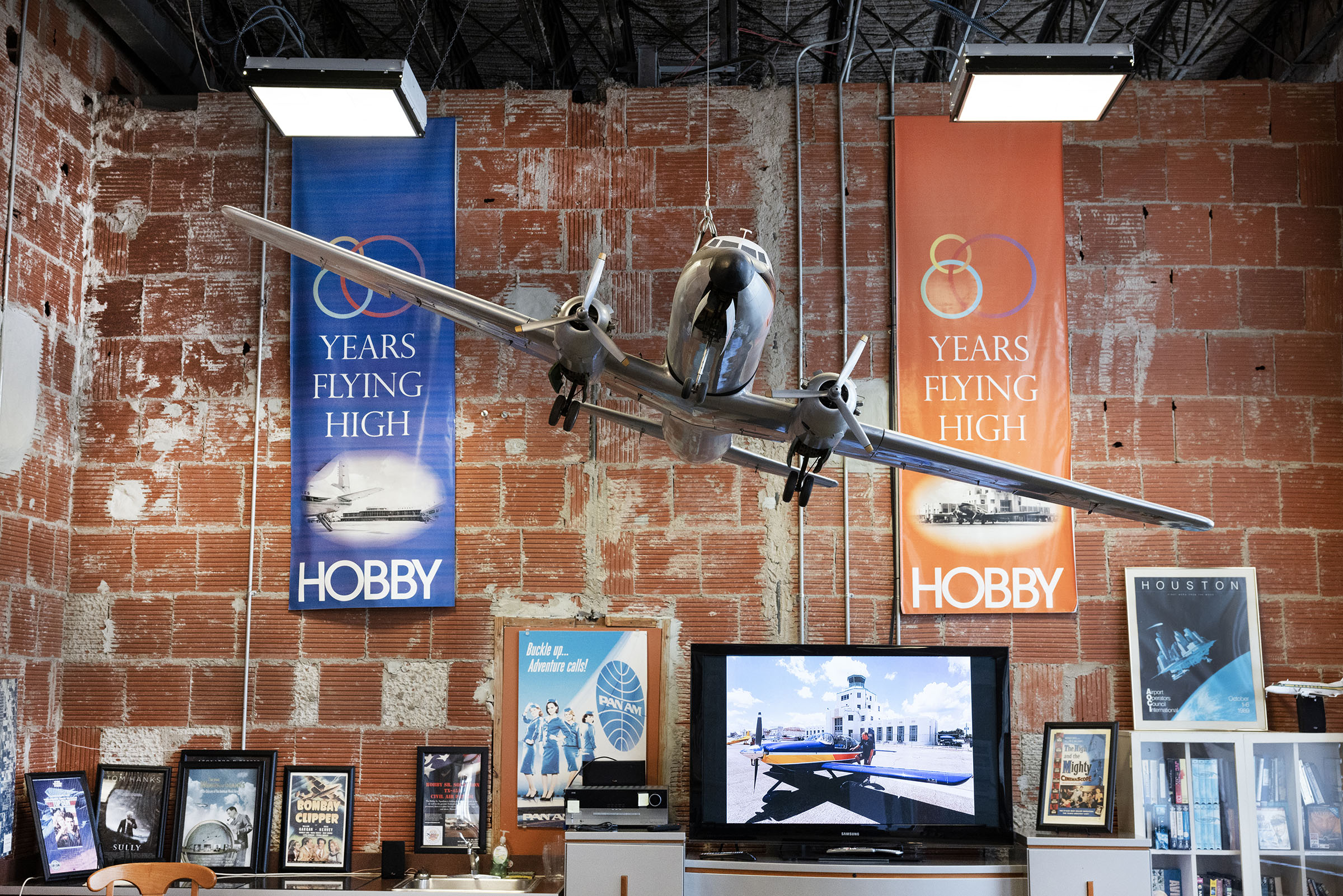 A glass display case contains photos, Pan Am posters, and banners printed with "50 Years Flying High" for Hobby Airport, and a model passenger jet hangs at a tilt. 