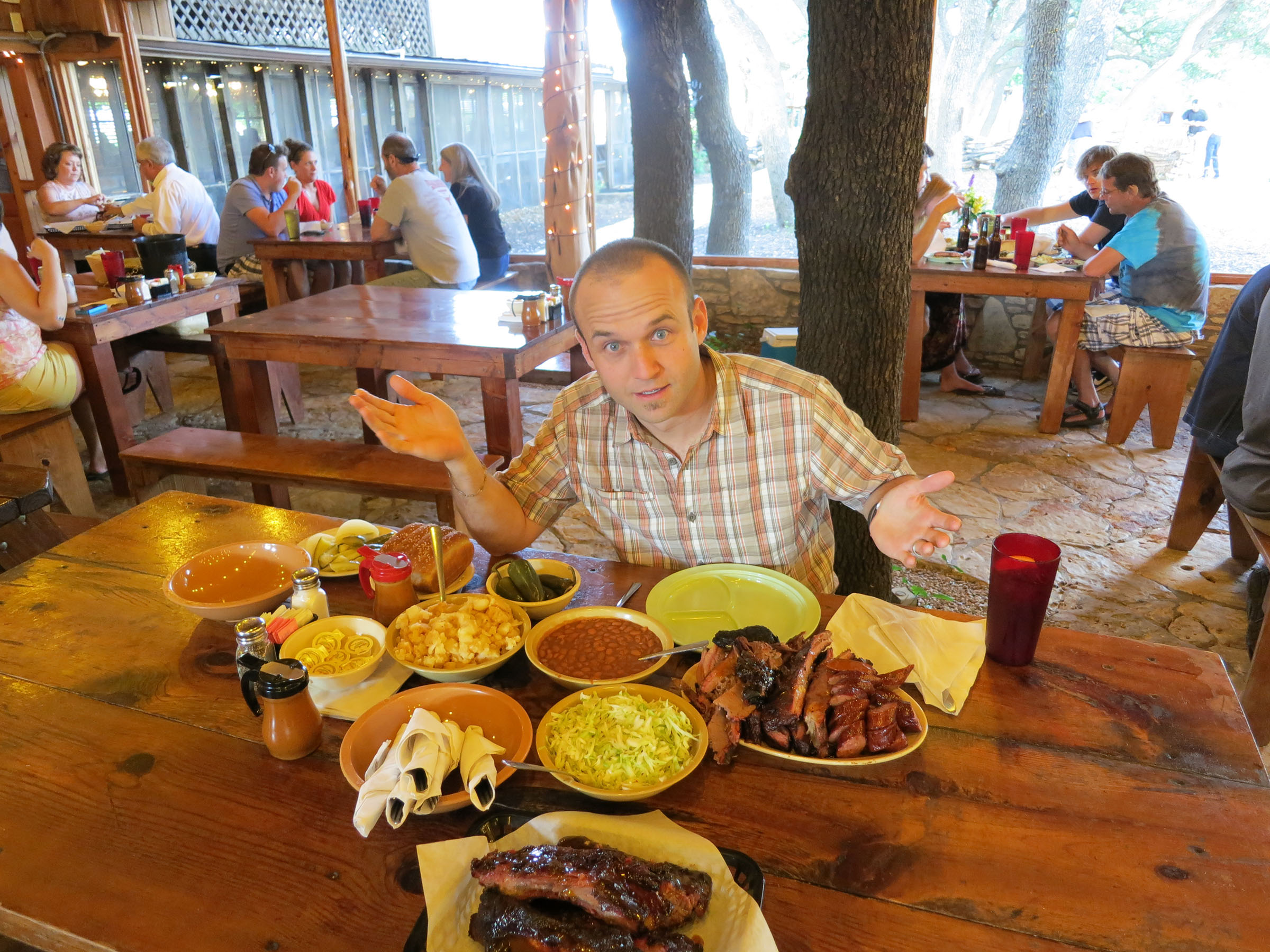 A man sits at a large table with numerous barbecue dishes and sides in an outdoor setting