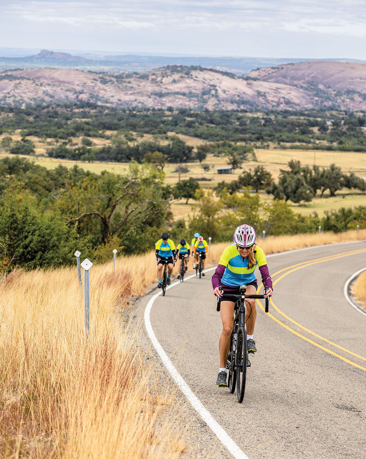 Three cyclists in bright green and blue shirts ascend an asphalt hill in the Texas hill country