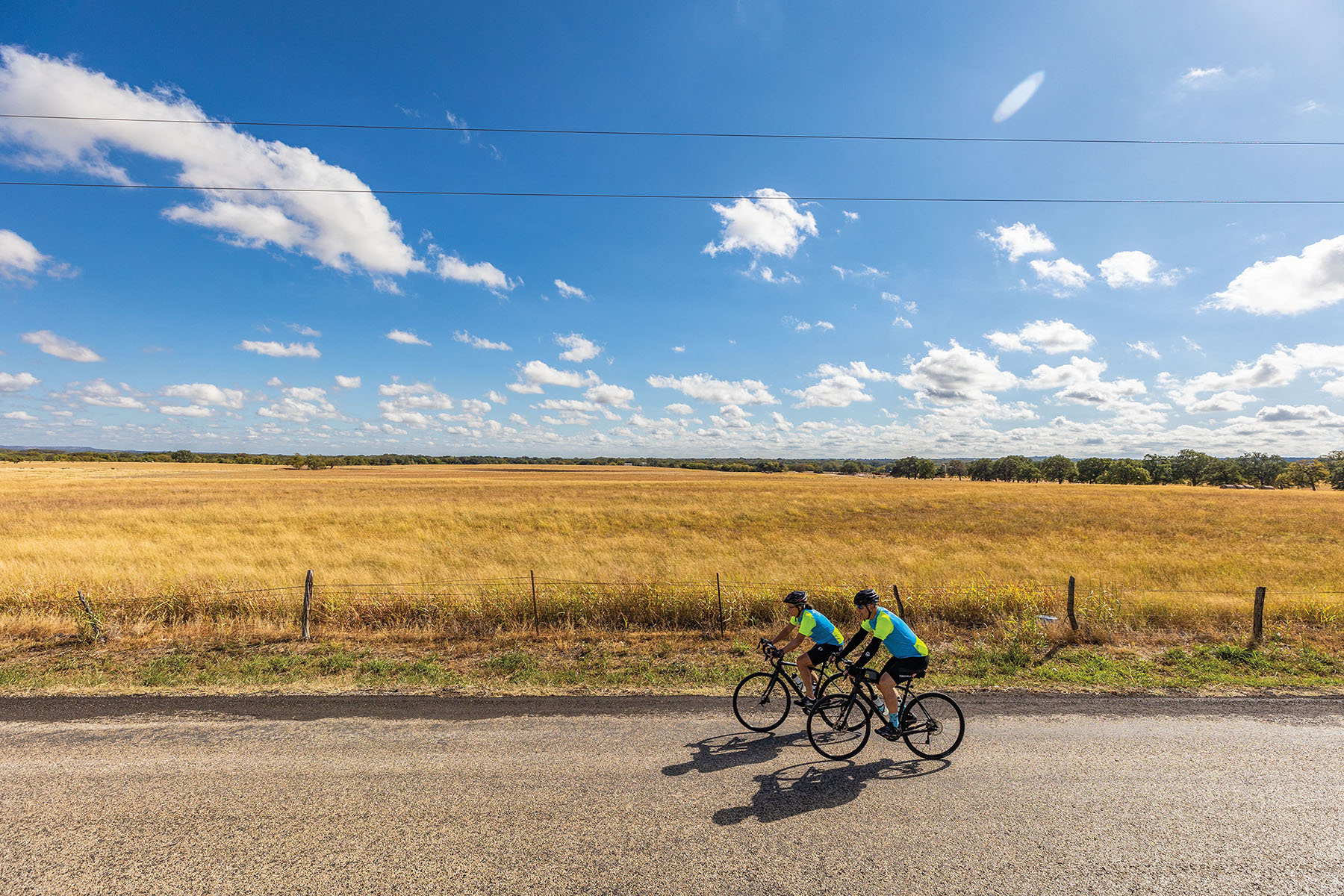Two bicyclists wearing blue and green shirts ride in front of a large field under blue sky