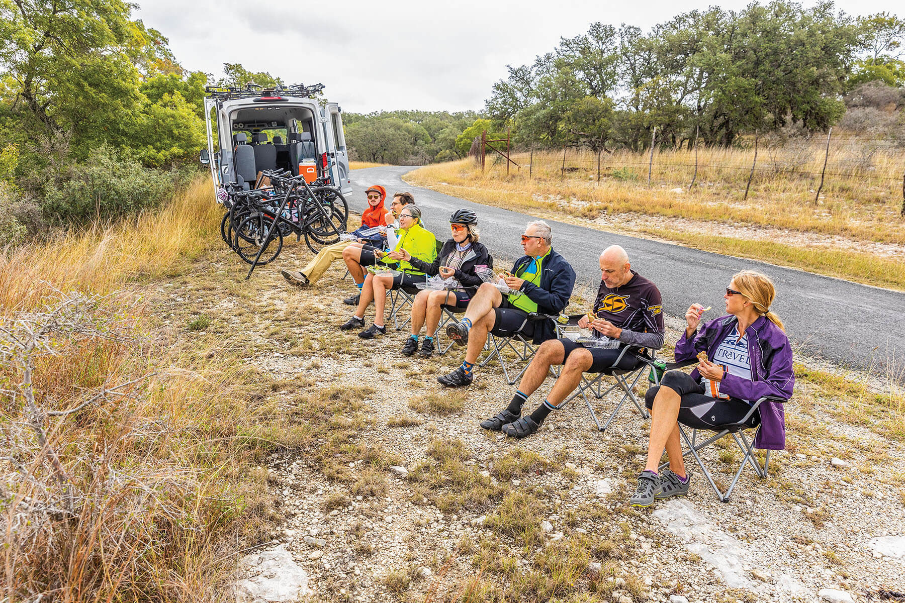 A group of cyclists in cycling gear sit on a curb next to a van