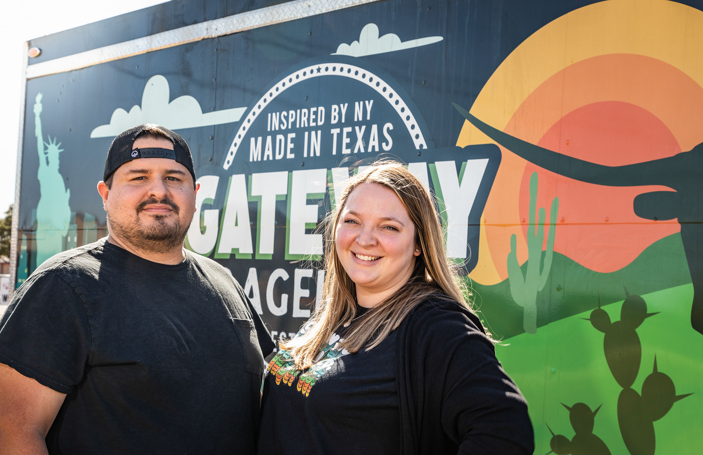 Two people in black shirts stand in front of a large mural that says "Inspired by NY, Made in Texas, Gateway Bagel Co"