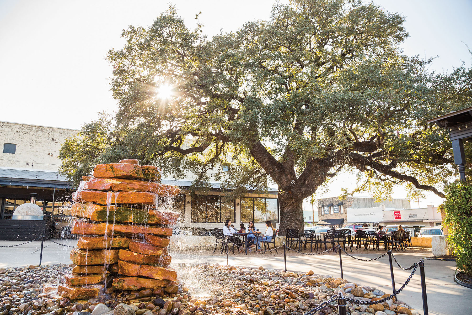 A stack of rocks and a fountain next to a large oak tree in the sunlight