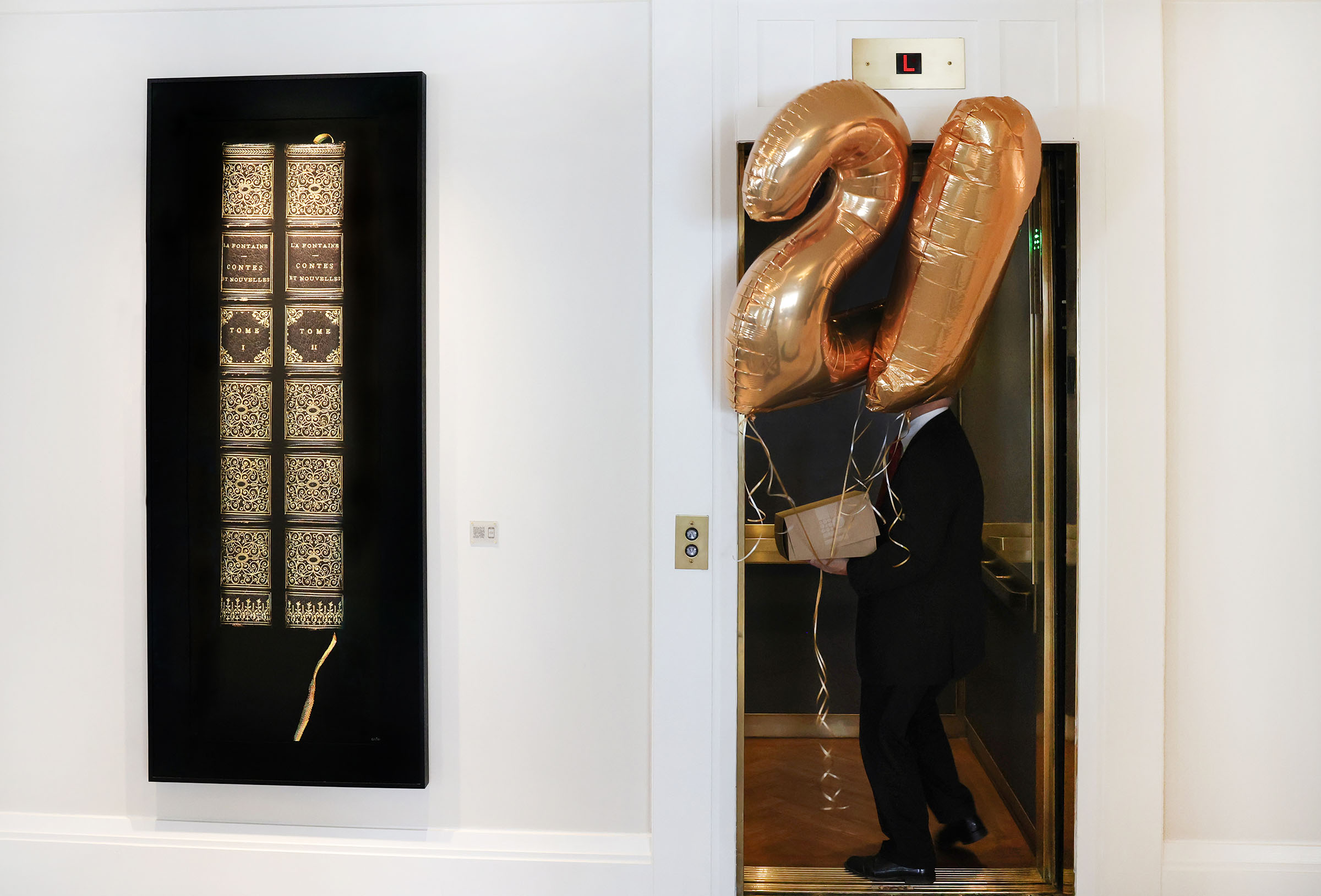 A person in formal attire carries large balloons into an elevator door next to a large painting of books