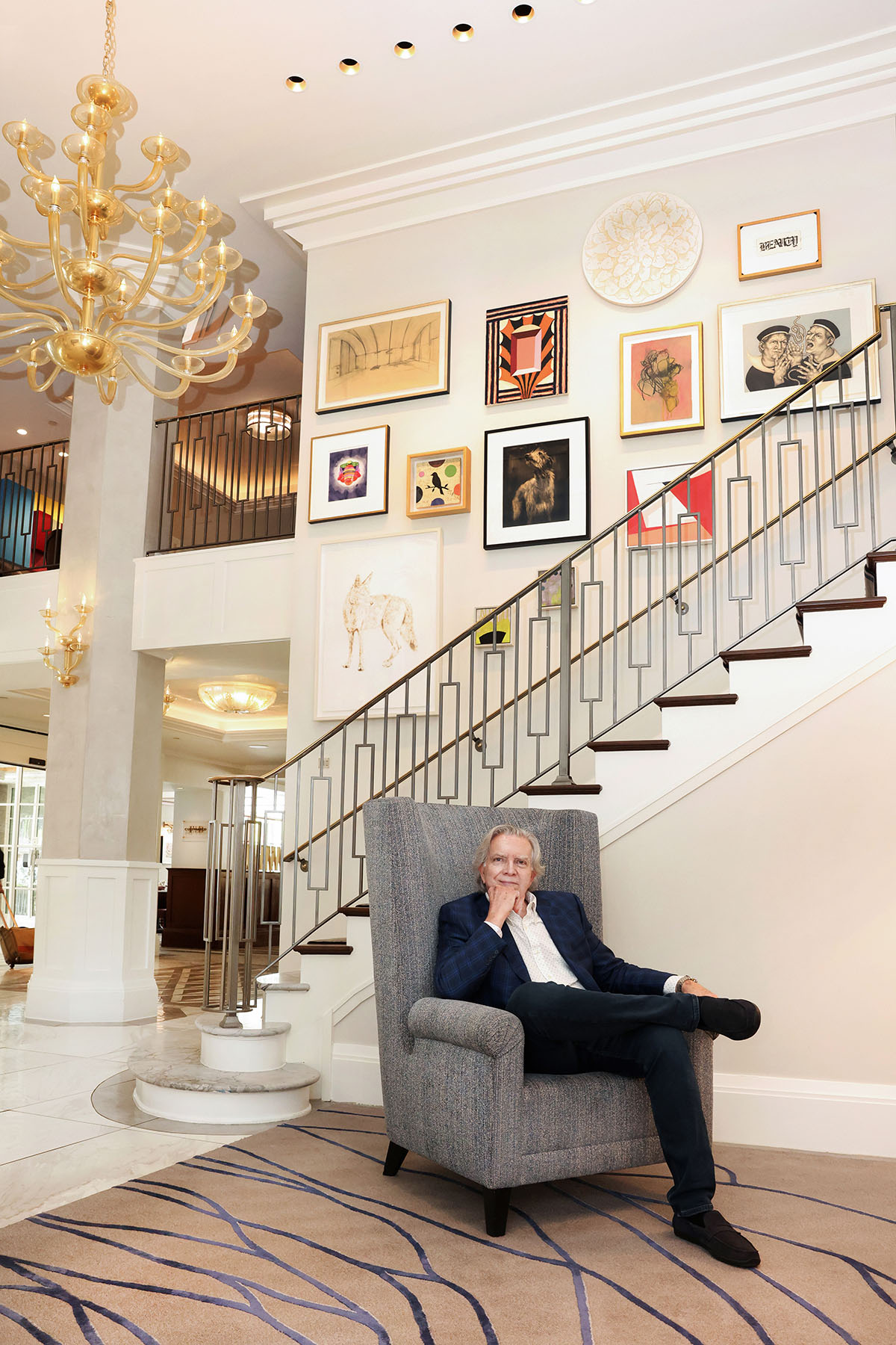 Jay Shinn sits in a gray armchair, a staircase and white wall covered in framed art behind him.