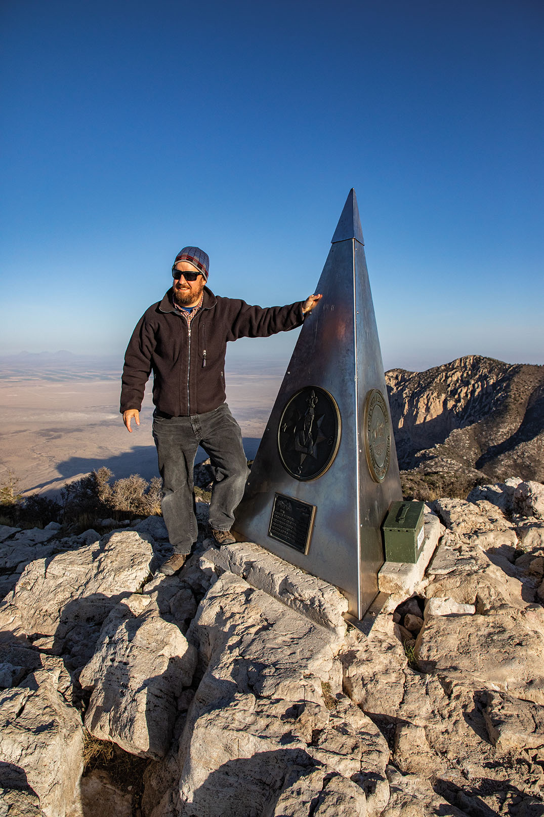 A man with sunglasses and a hat stands in front of an obelisk that is on top of a mountain.