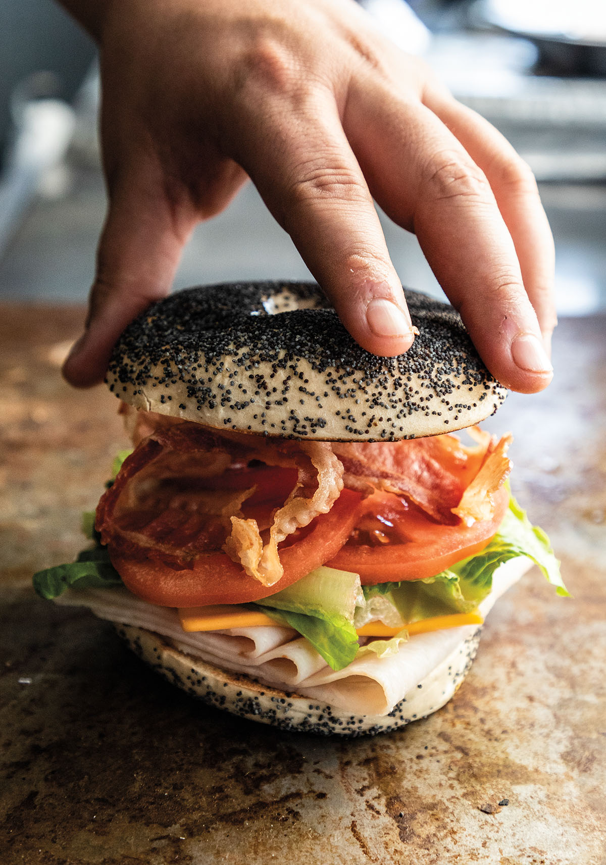 A hand places a poppy seed top on a stacked bagel with turkey, avocado, tomato, and bacon