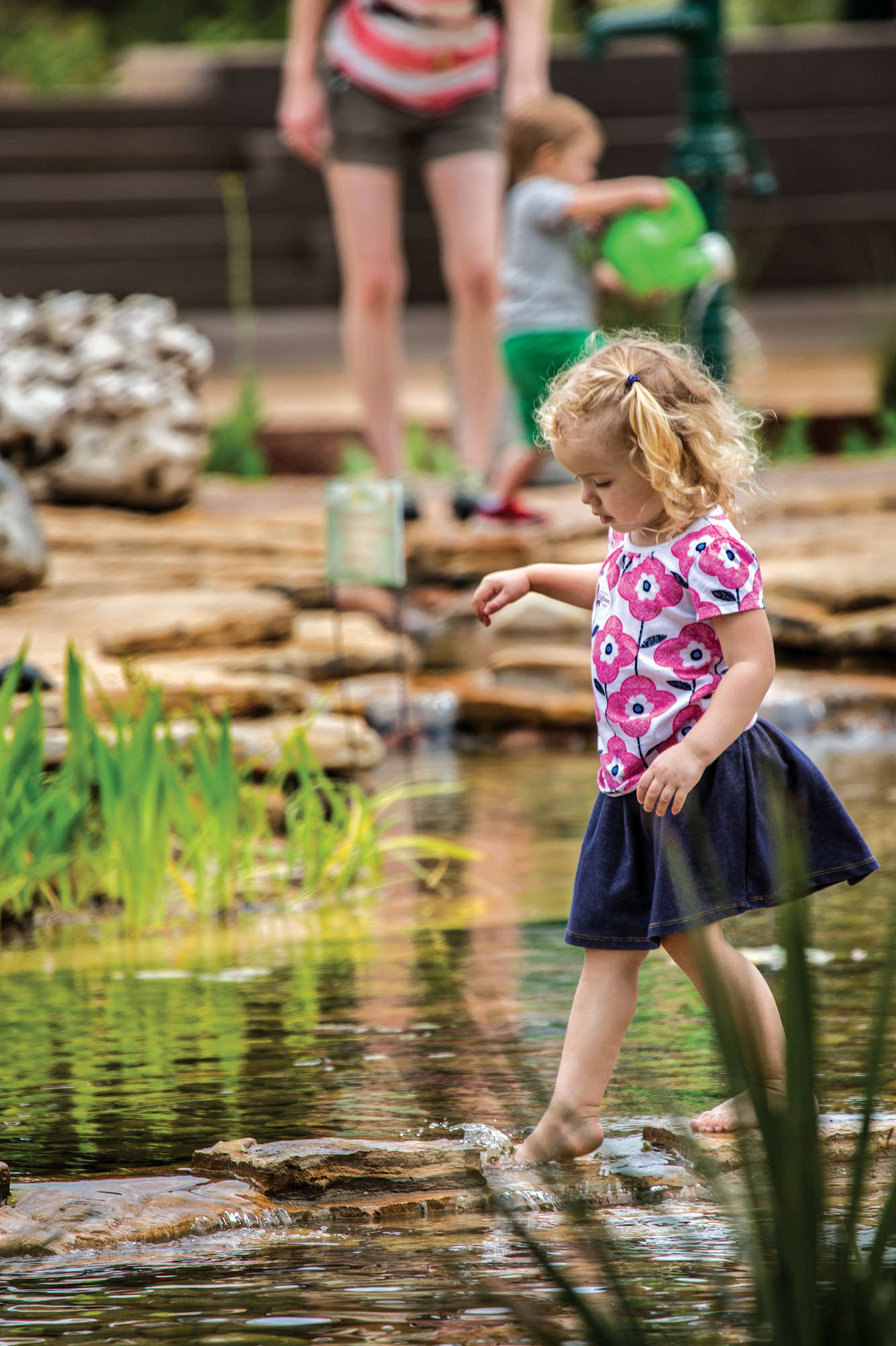 A child dips her toes into a pond at the Lady Bird Johnson Wildflower Center's Family Garden that recreates the Paluxy River ecosystem in which Dinosaur Tracks were found in Glen Rose. A woman and a younger boy stand in the background