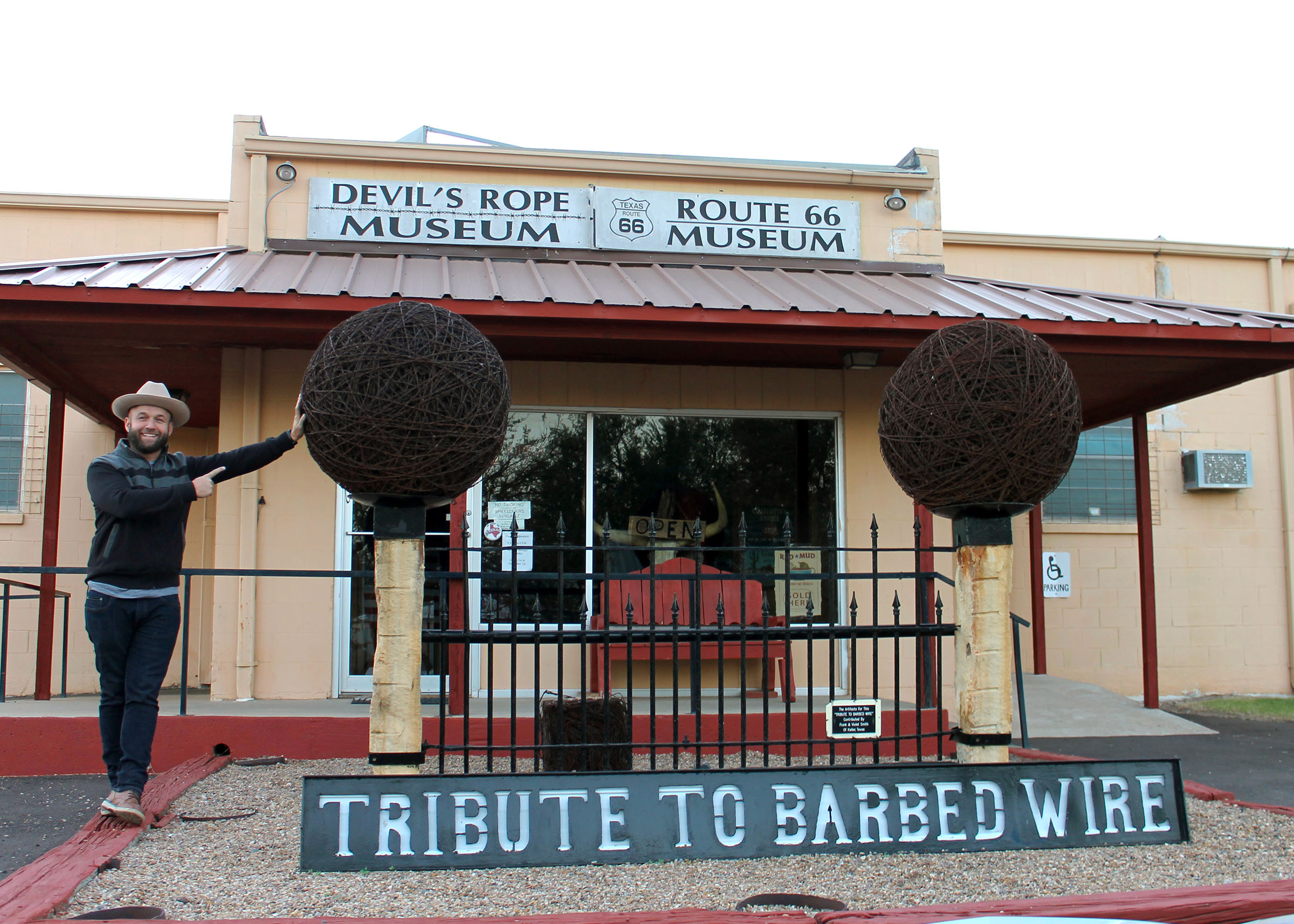A man in a cowboy hat stands outside of a building with a sign reading "Devil's Rope Museum: Tribute to Barbed Wire"