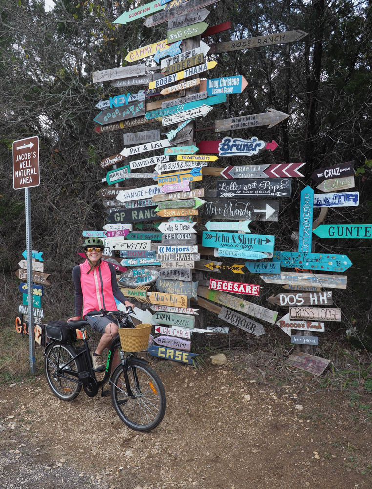 A woman in a pink vest stands next to a collection of bright painted signs 