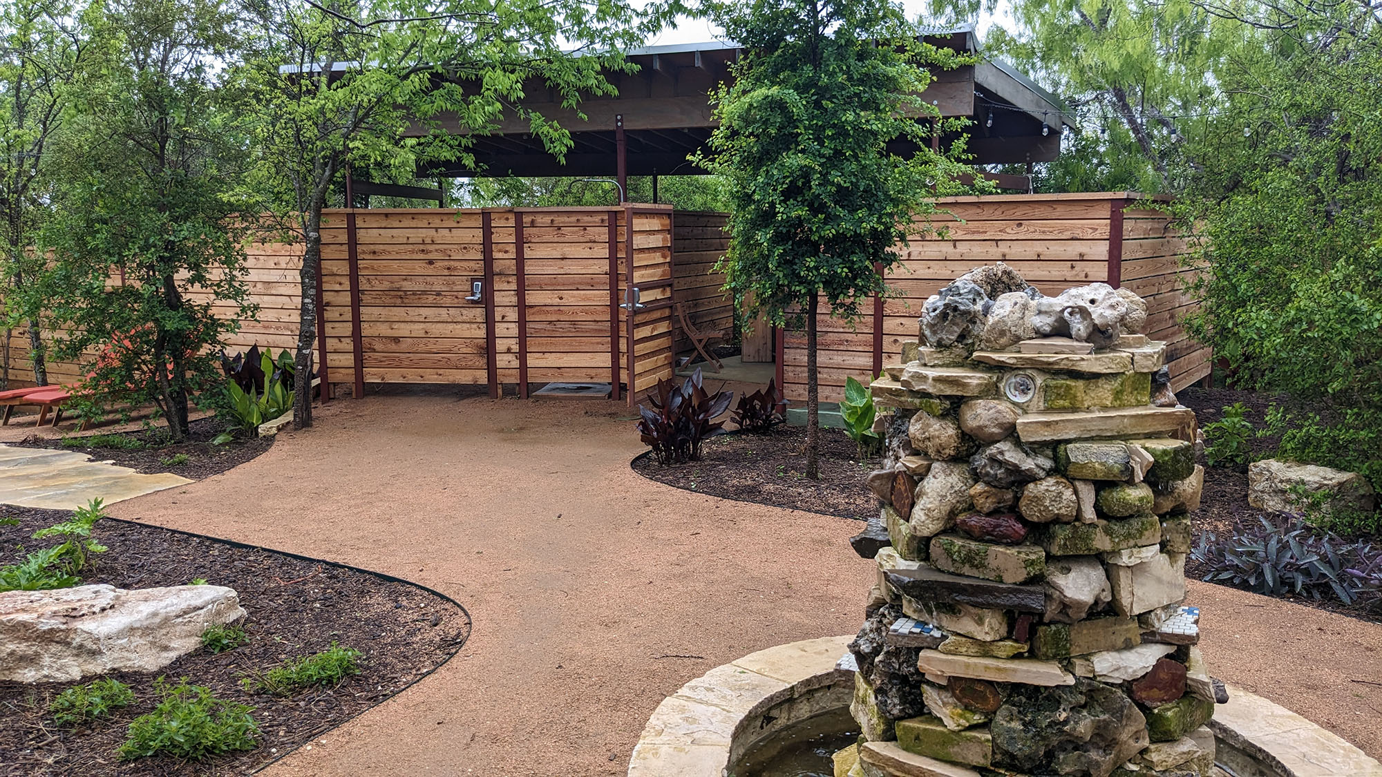 A winding path with small gardens on the left and right leads to the entrance, where horizontal wooden fencing hides the interior. A fountain made of slabs and rocks is in the foreground. 