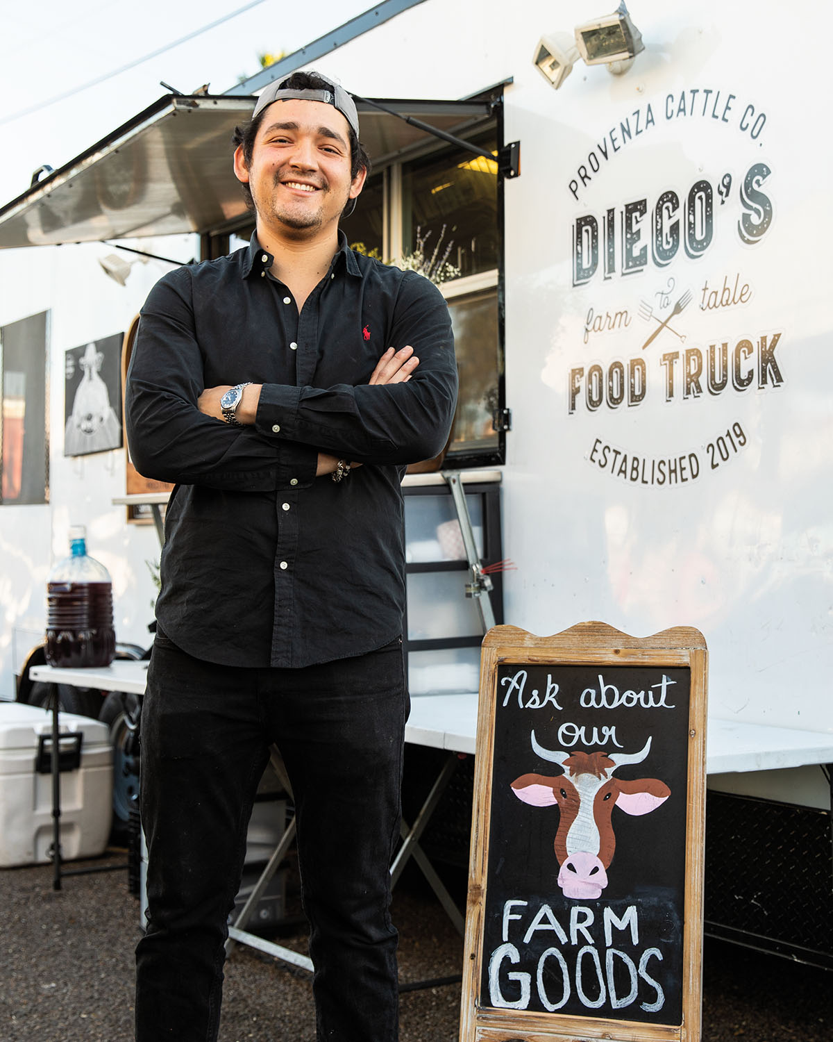 A man in a black button-up shirt stands with his arms crossed in front of a white food truck