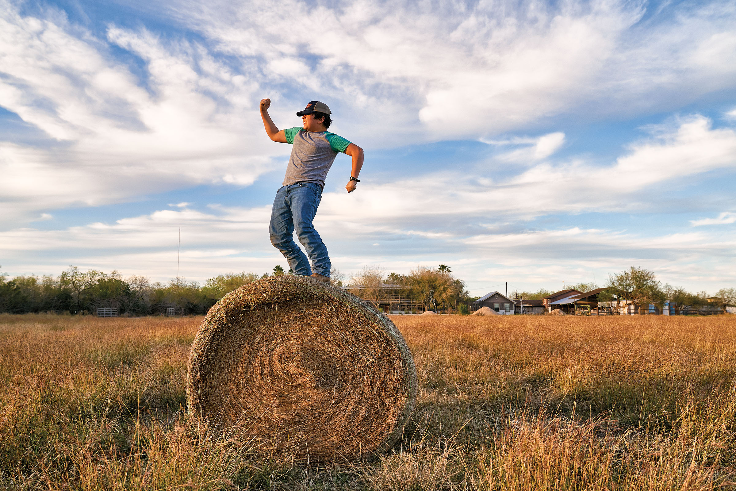 A young man in a t-shirt and jeans stands on a large circular haybale