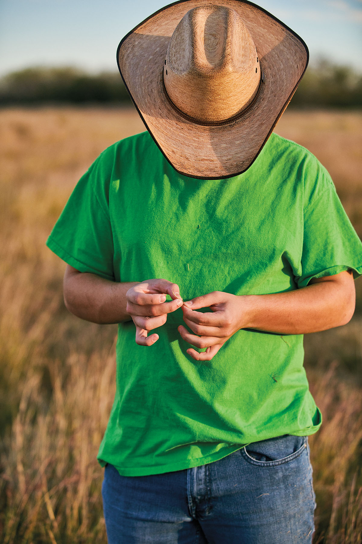 A young person in a green shirt and cowboy hat poses with his head down in a grassy field