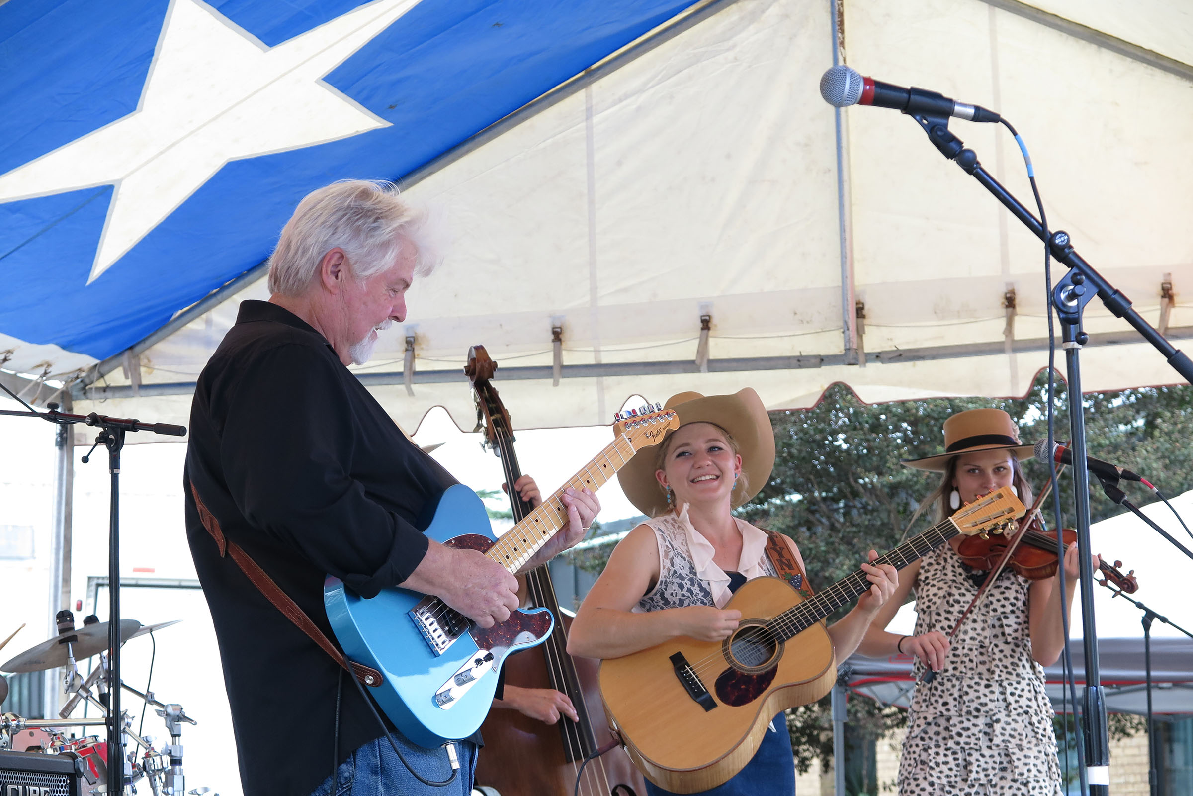 Three people play guitars and fiddle on a stage underneath a large Texas flag tent