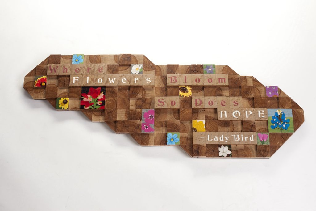 A slab of wood has Scrabble-like tiles that are arranged with a quote from Lady Bird Johnson: "Where Wildflowers Bloom, So Does Hope." Other tiles contain painted wildflowers. 