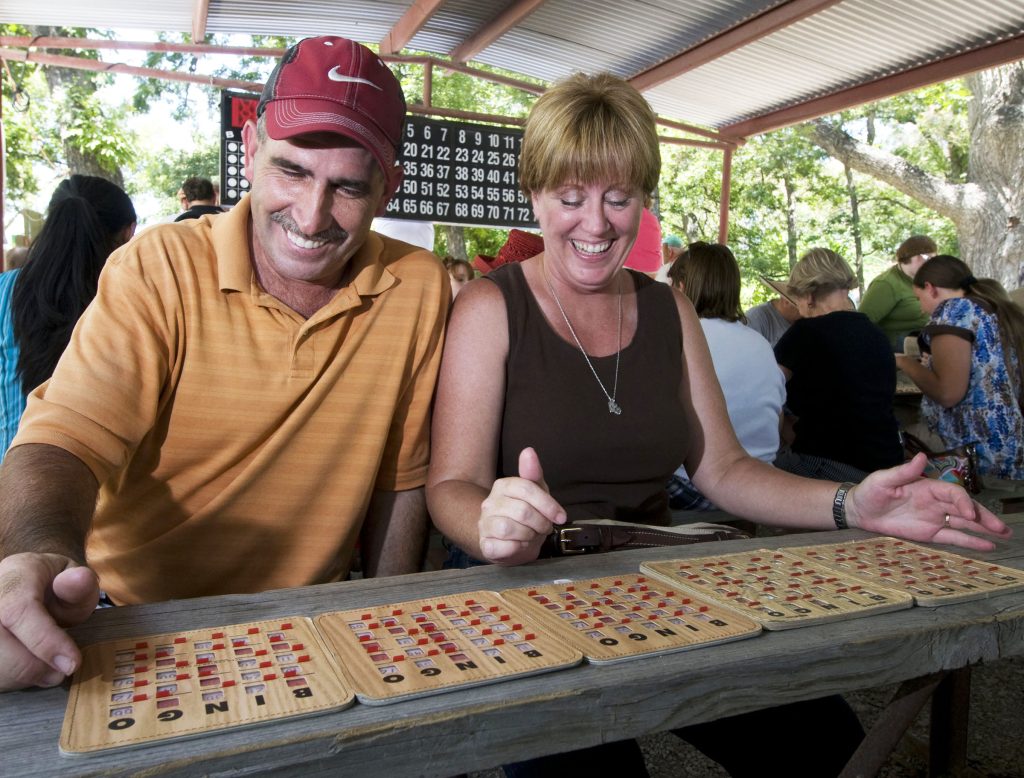 A man and woman sit in front of five bingo cards with a large bingo board in the background
