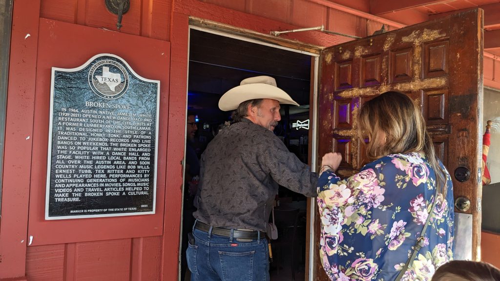 The historical marker is attached to the wall next to the front door of the honky-tonk. A man wearing a white cowboy hat and Western-style clothes has the door open as he enters the venue while a women wearing a flow-y floral top enters after him. 