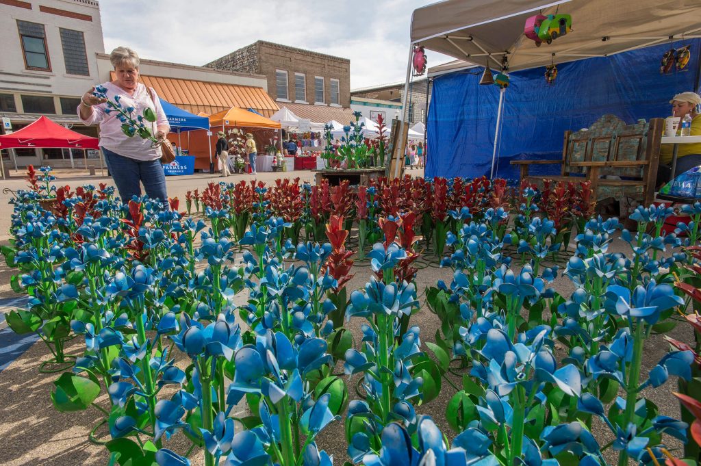 A woman holds a large metal bluebonnet among several dozen other metal bluebonnets in blue and red during an outdoor festival