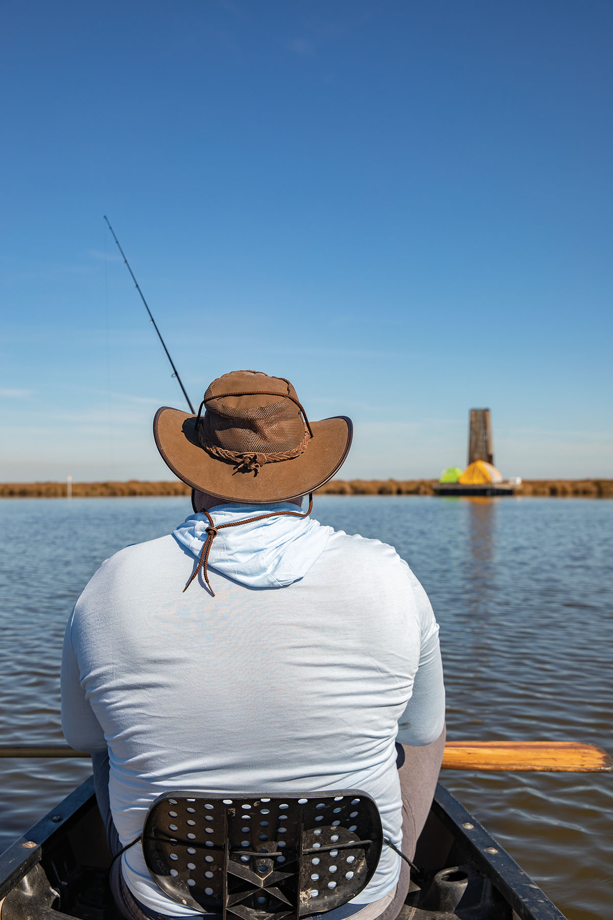 A person in a light blue shirt and brown hat casts a reel over a flat blue body of water with the floating tenpt pad in the distance