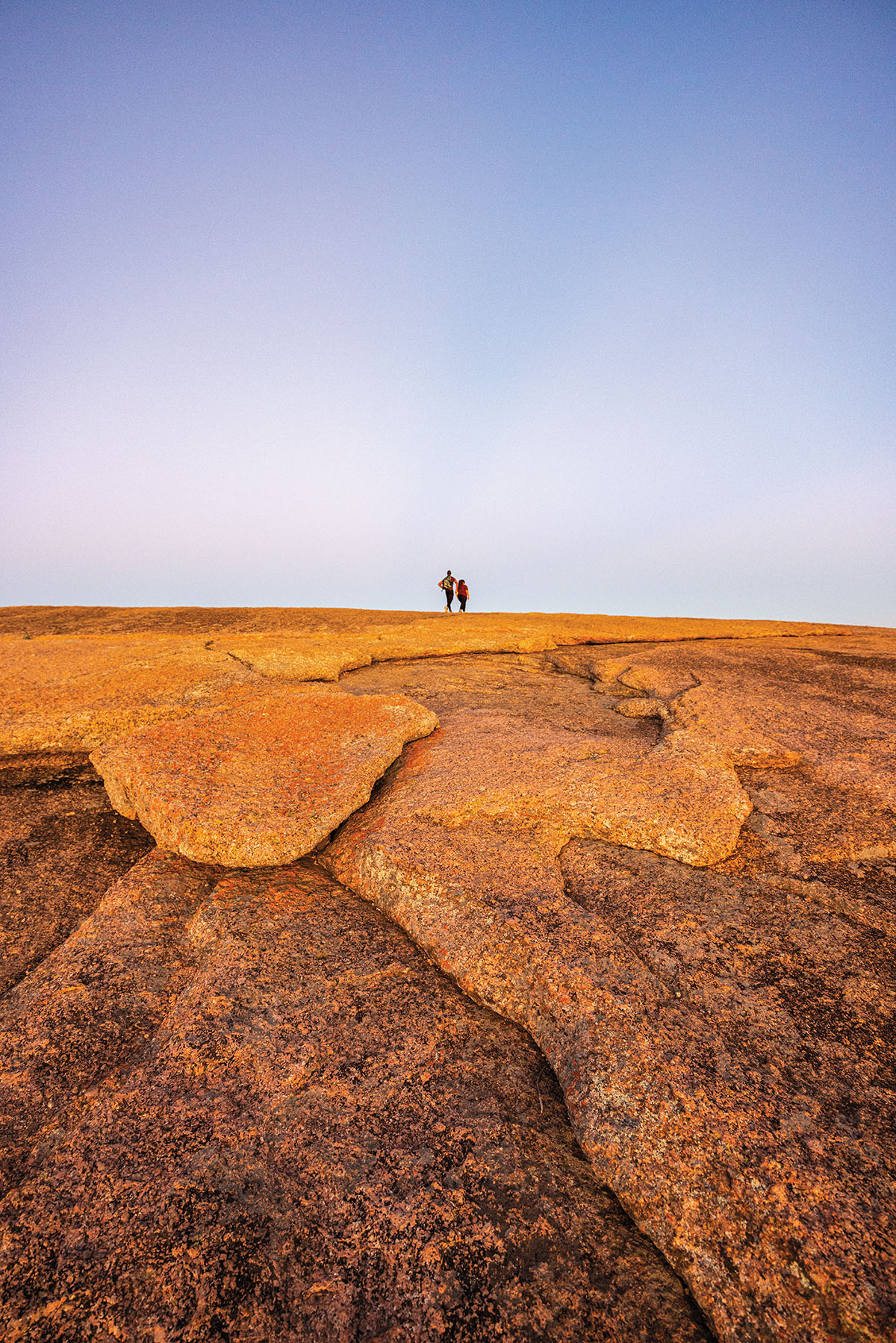Two hikers stand on top of the golden-brown surface of Enchanted Rock, beneath a purple gradient sky