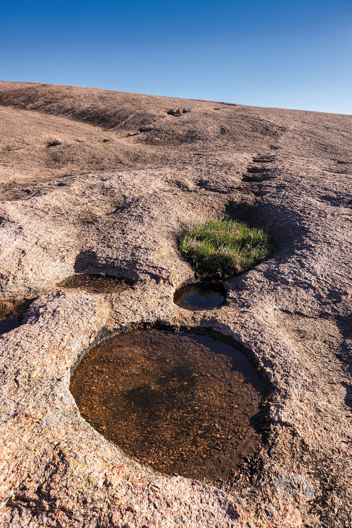Small pools of brown water cover the dark stone landscape under blue sky at Enchanted Rock