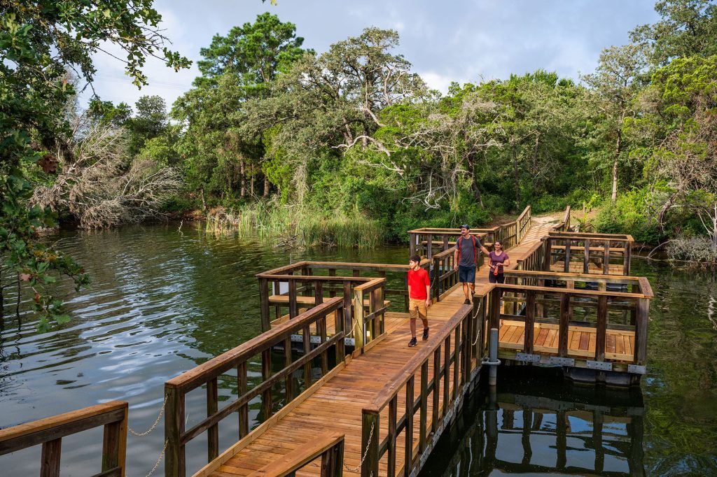 A man and woman and an older boy walk across a wooden boardwalk with viewing areas along Lake Bistro. Lush trees and other vegetation are in the background. 