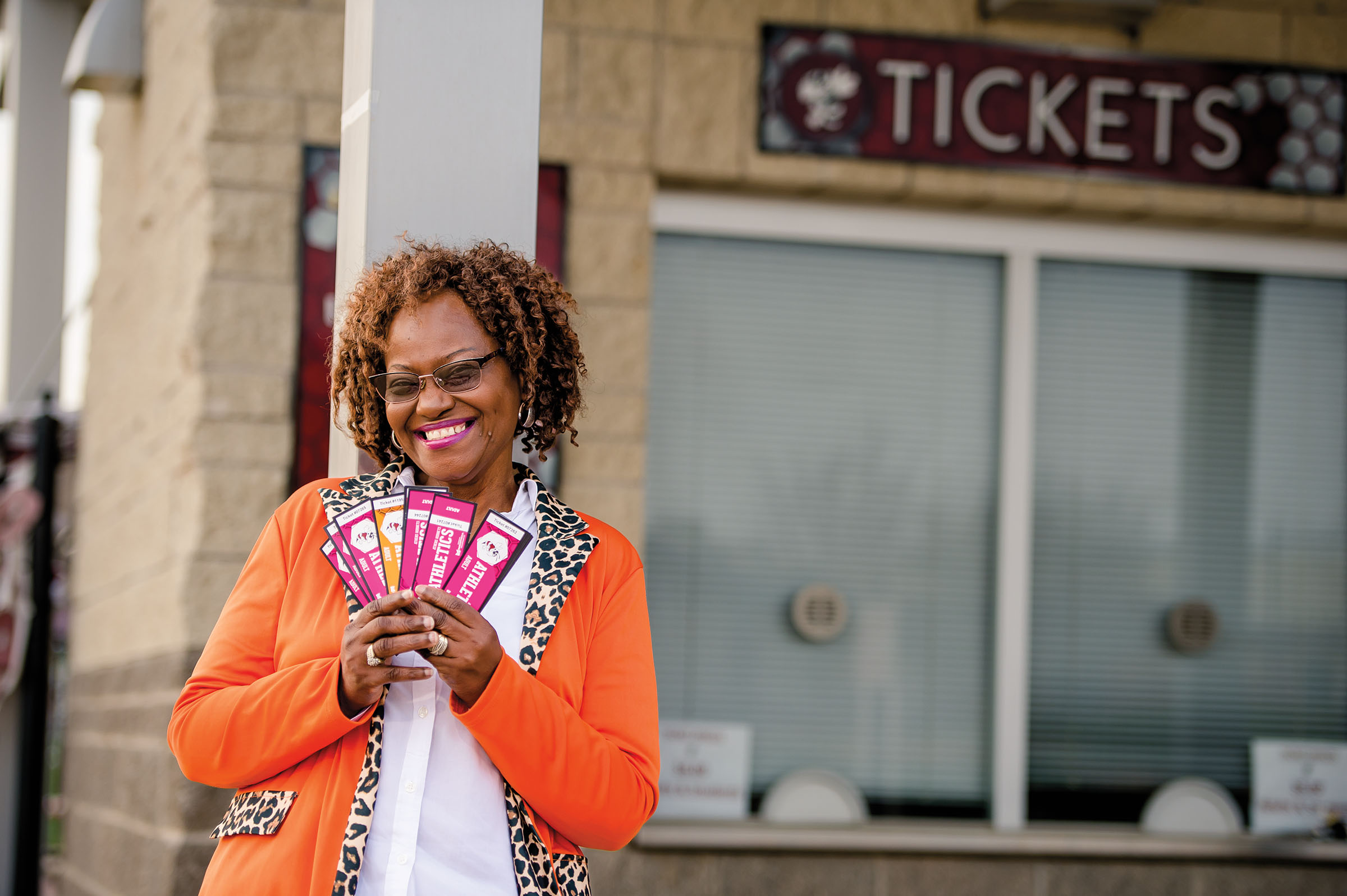 A woman in a bright orange blazer holds a handful of brightly-colored sports tickets