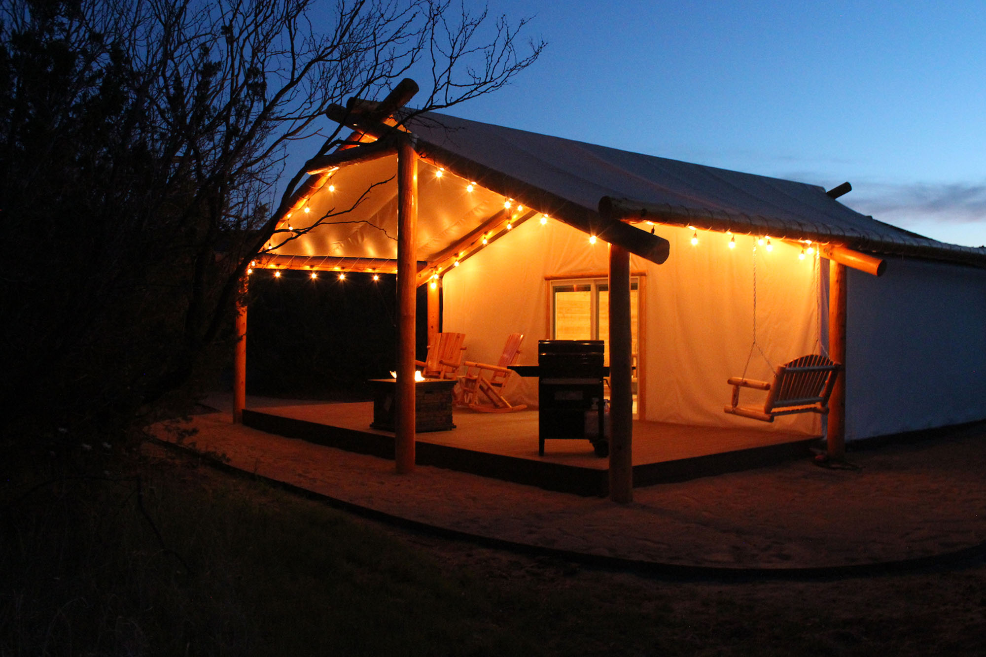 A nighttime shot of one of the glamping cabins shows lit string lights, a grill, and seating on a large porch. 