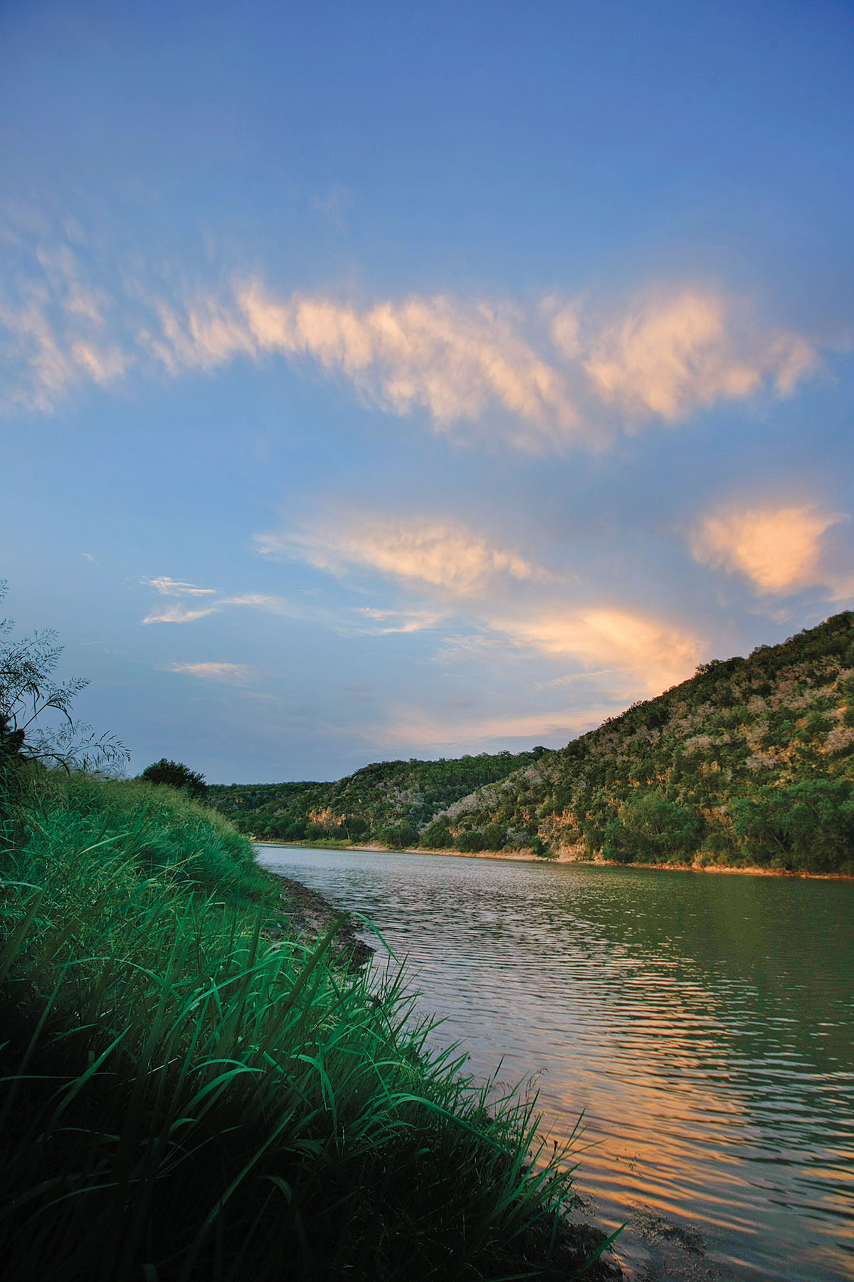 Whispy clouds on a blue sky above deep water under rolling green hills at Colorado Bend State Park