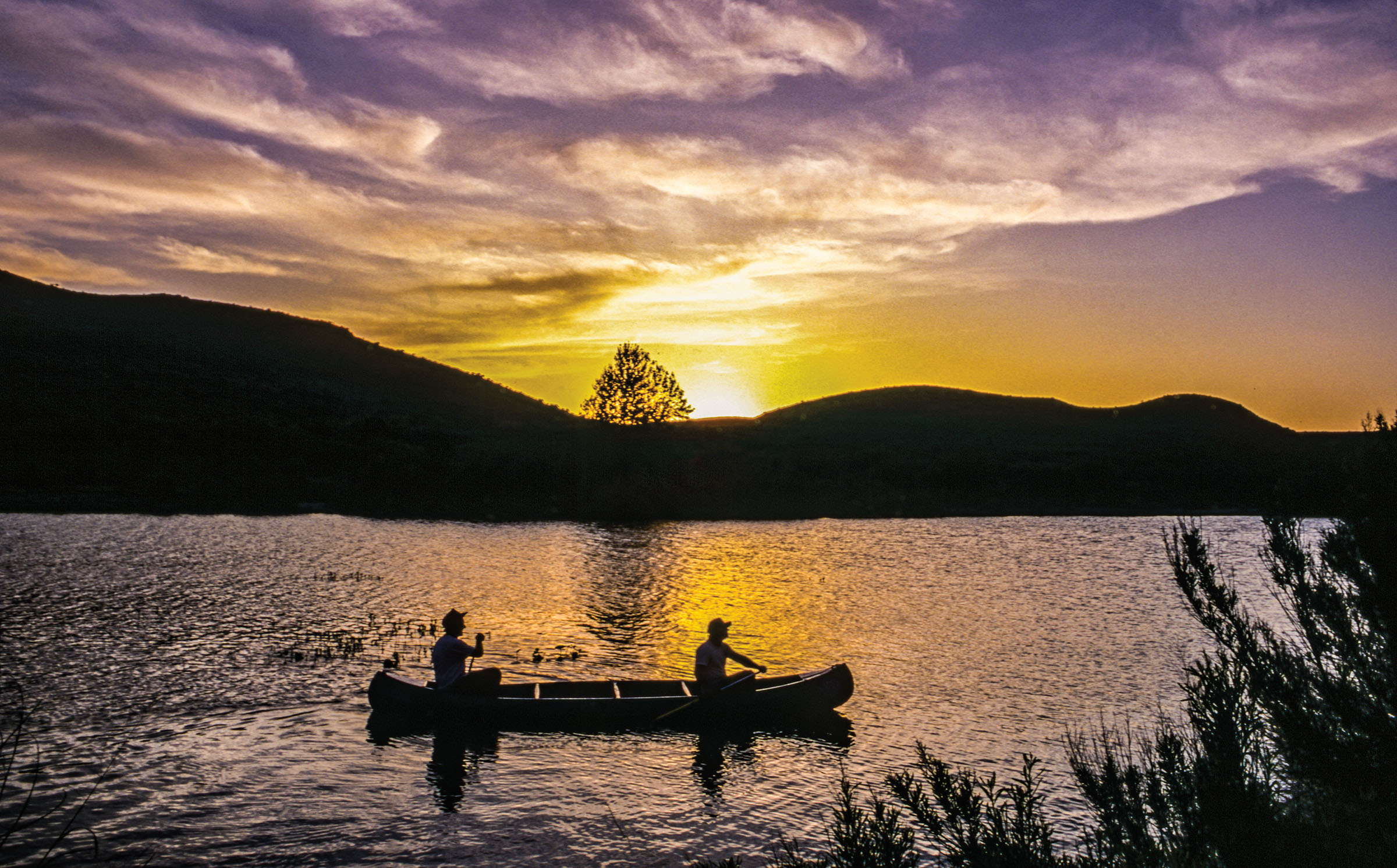 Two people paddle a canoe in front of a sunset over dark hills