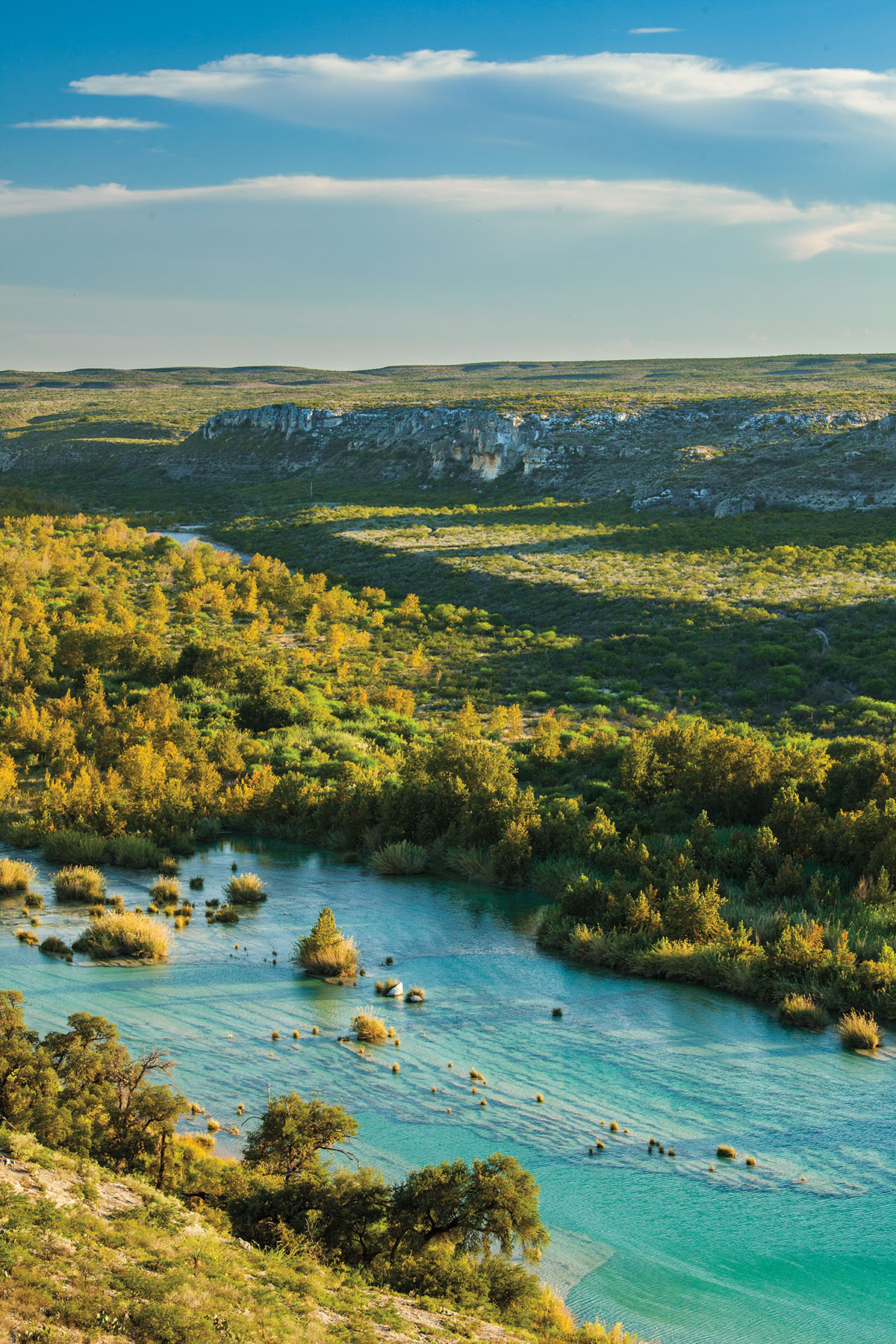 Bright blue-green water flows through a vast green hill country expanse