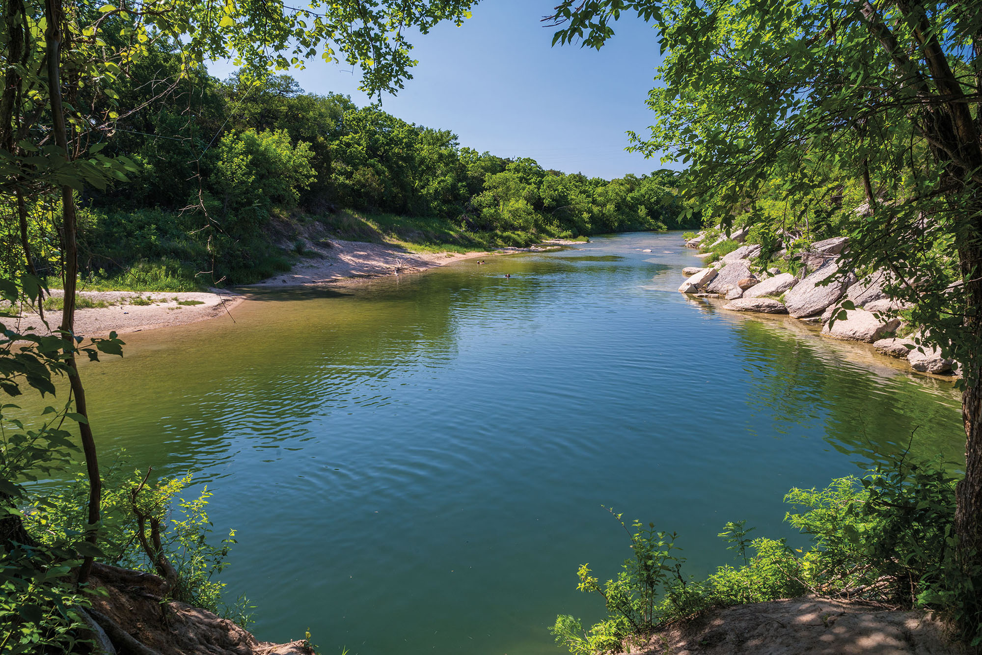 A view through green trees of pristine blue water under blue sky with limestone rocks on the shore