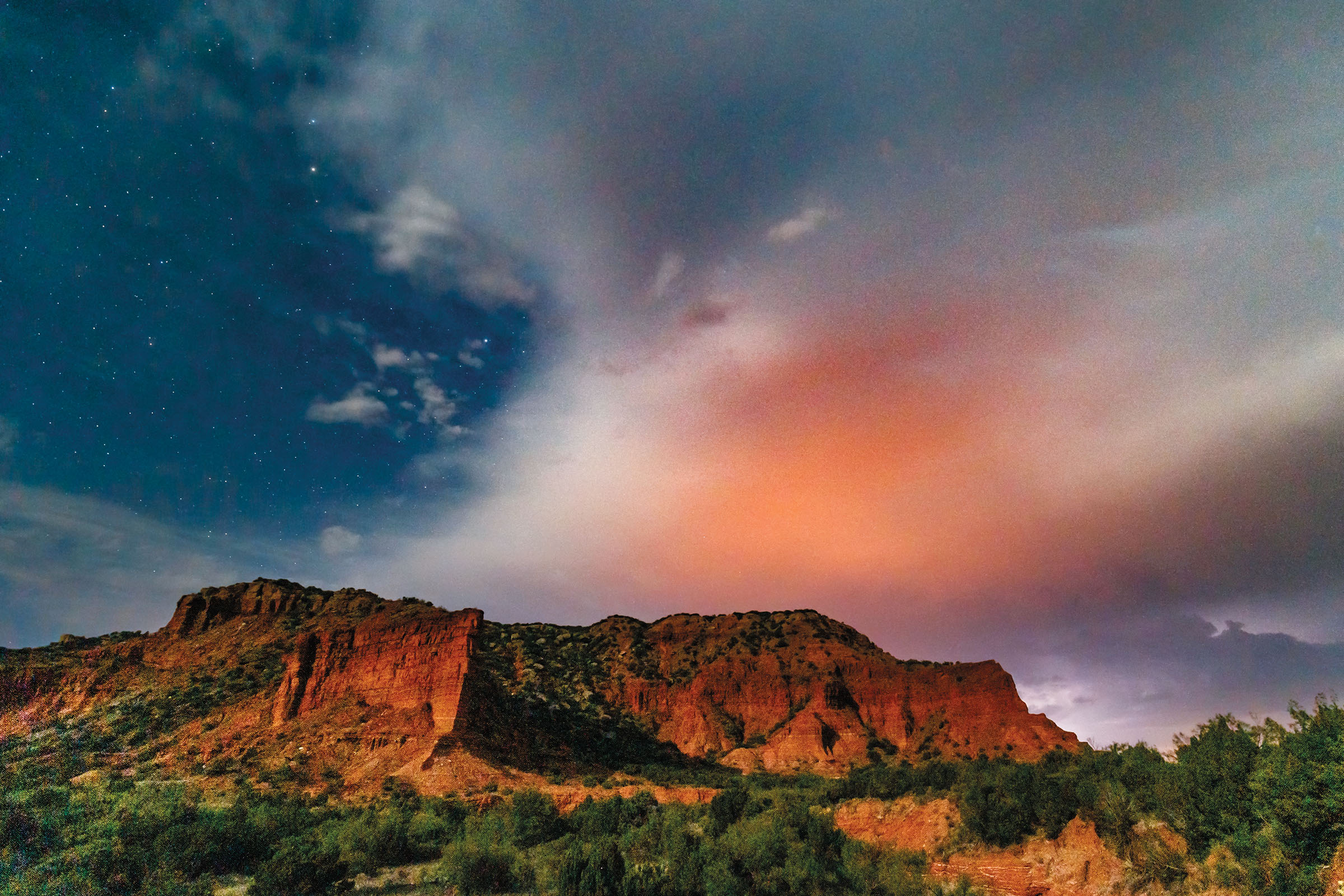 A large red rock outcropping under a large cloud with a pink tint on a dark sky
