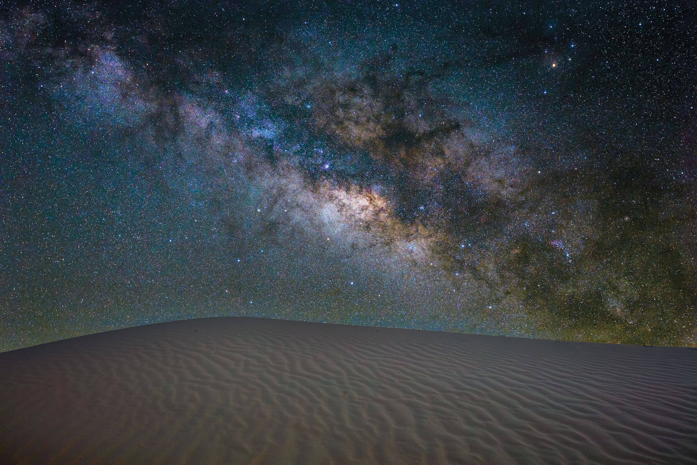 The Milky Way galaxy is visible in purple and blue tones, along with thousands of stars, above a sandy floor